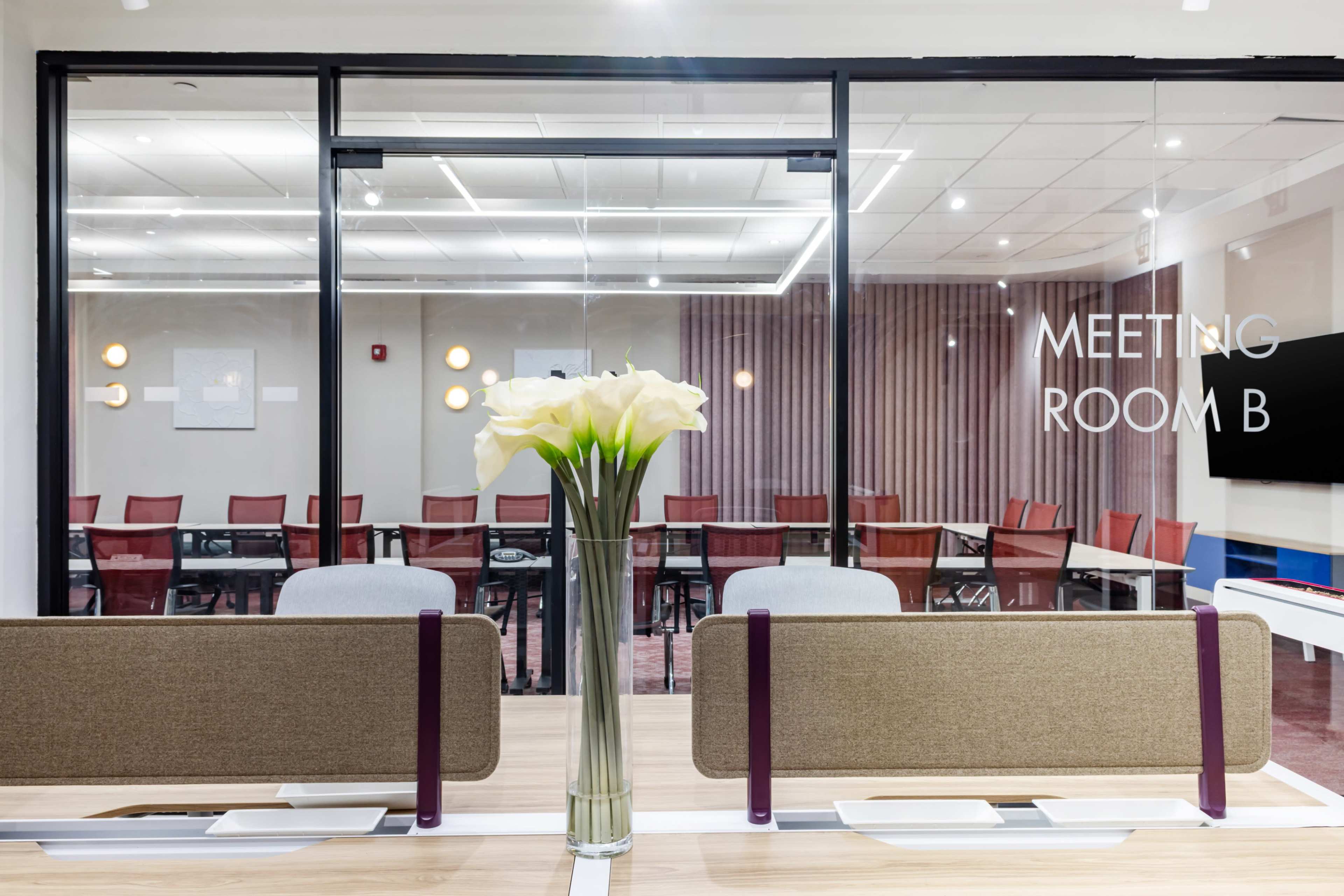 A conference room is visible through glass walls, featuring a table with chairs, and a vase of white flowers in the foreground.