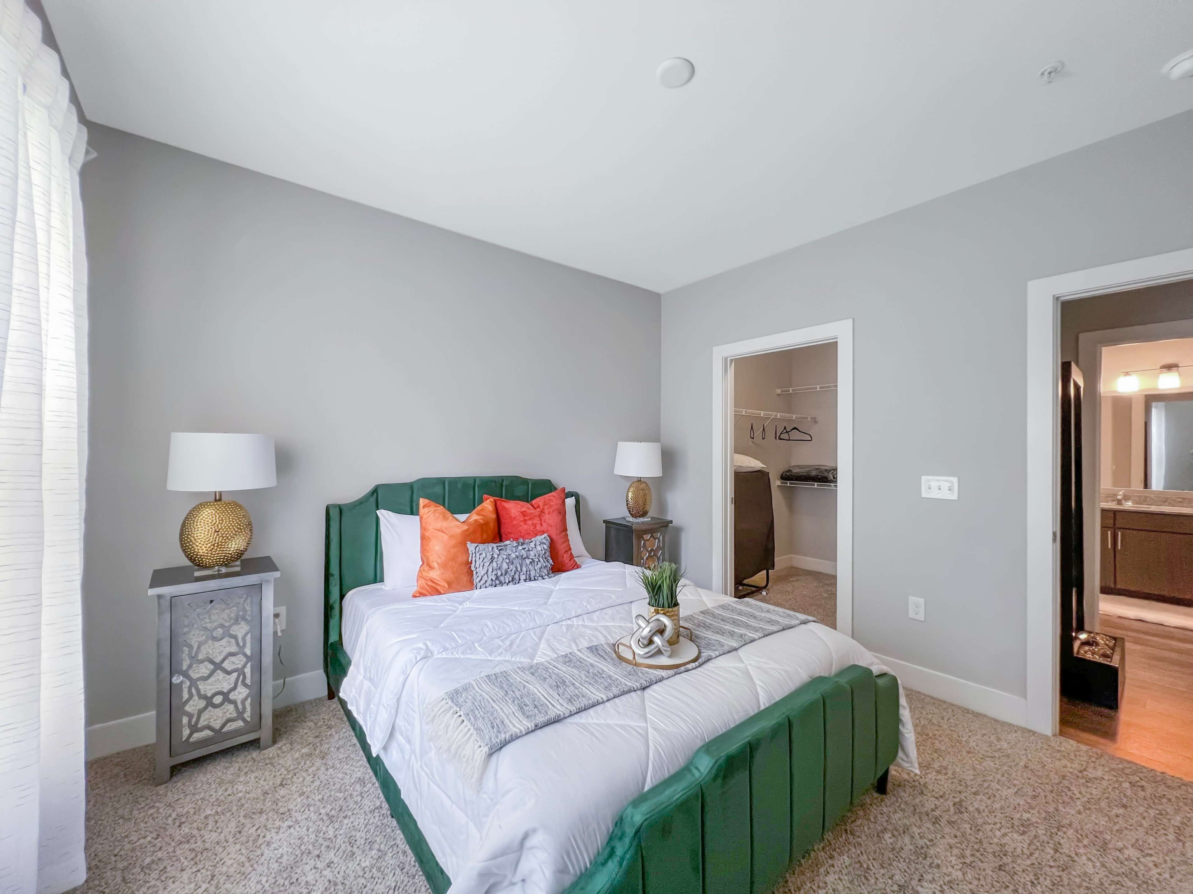 A neatly arranged bedroom features a green upholstered bed with decorative pillows, two bedside lamps, and an attached bathroom visible through a doorway.
