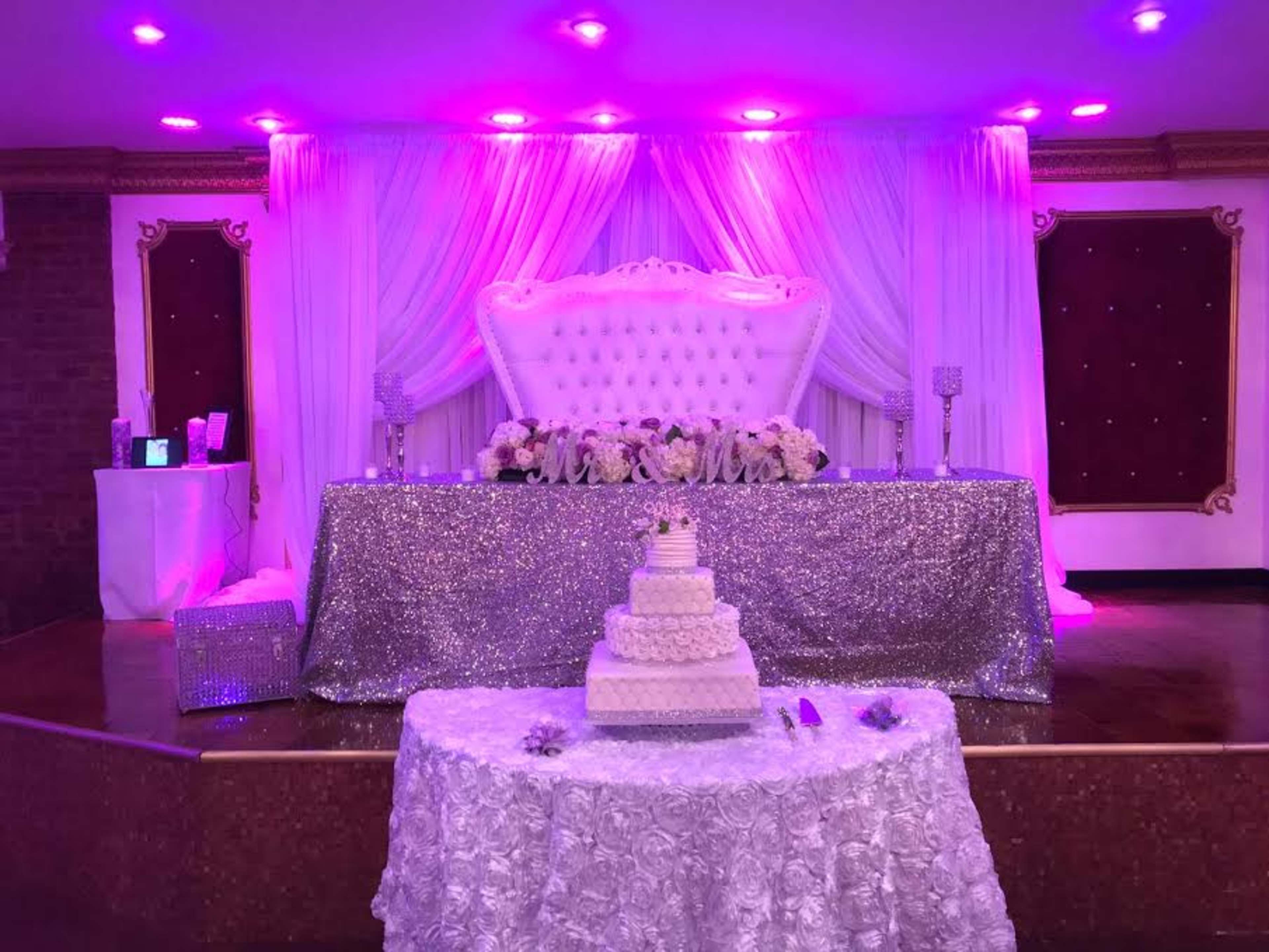 The image shows a decorated event space with a white and silver cake on a lace tablecloth in front of a lavish backdrop featuring a large tufted white settee and draped fabric illuminated by purple lighting.