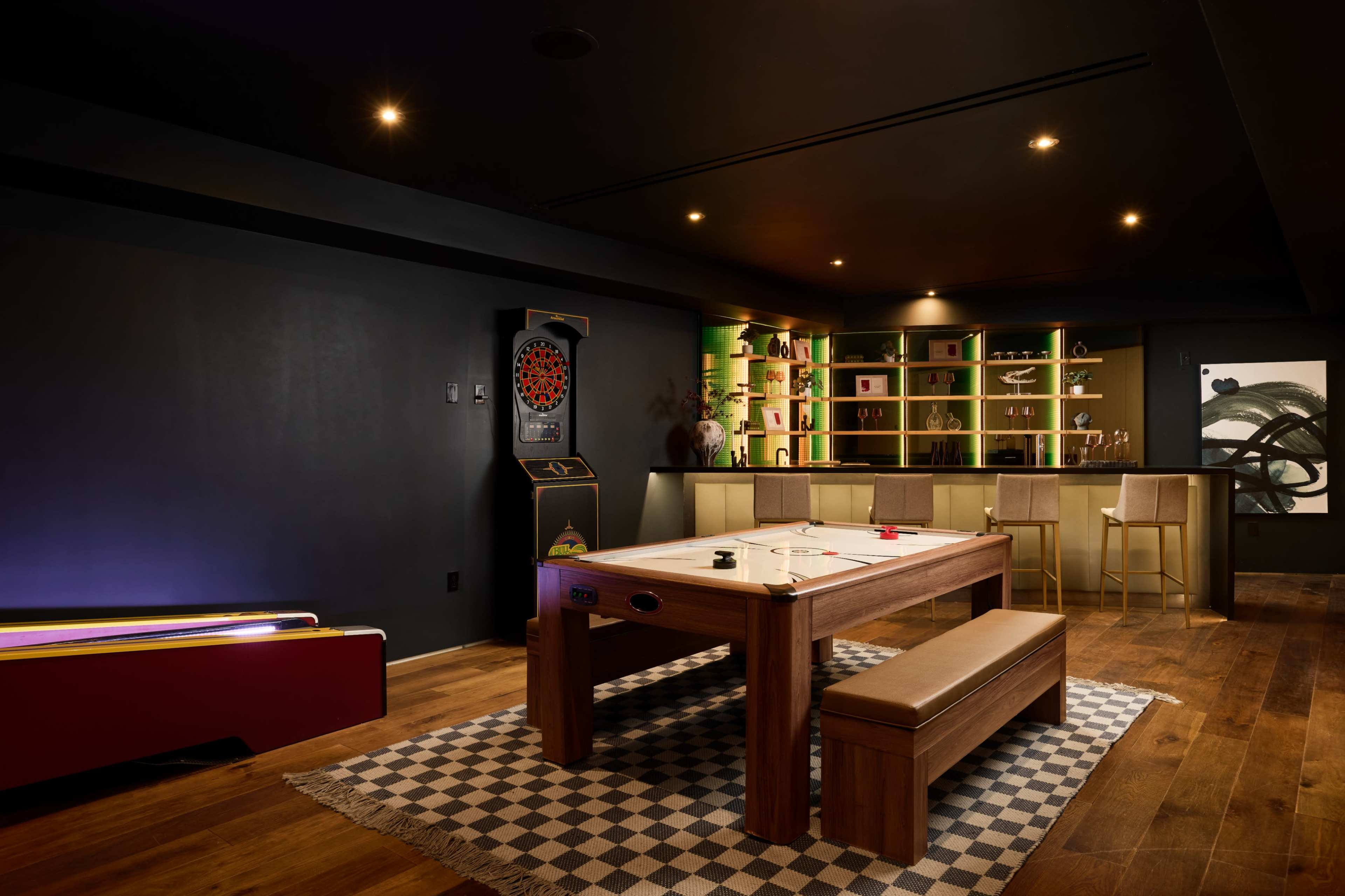 The image shows a modern game room featuring a wooden air hockey table, a bar with shelves displaying various items, and a dartboard on the wall, all illuminated by recessed lighting.