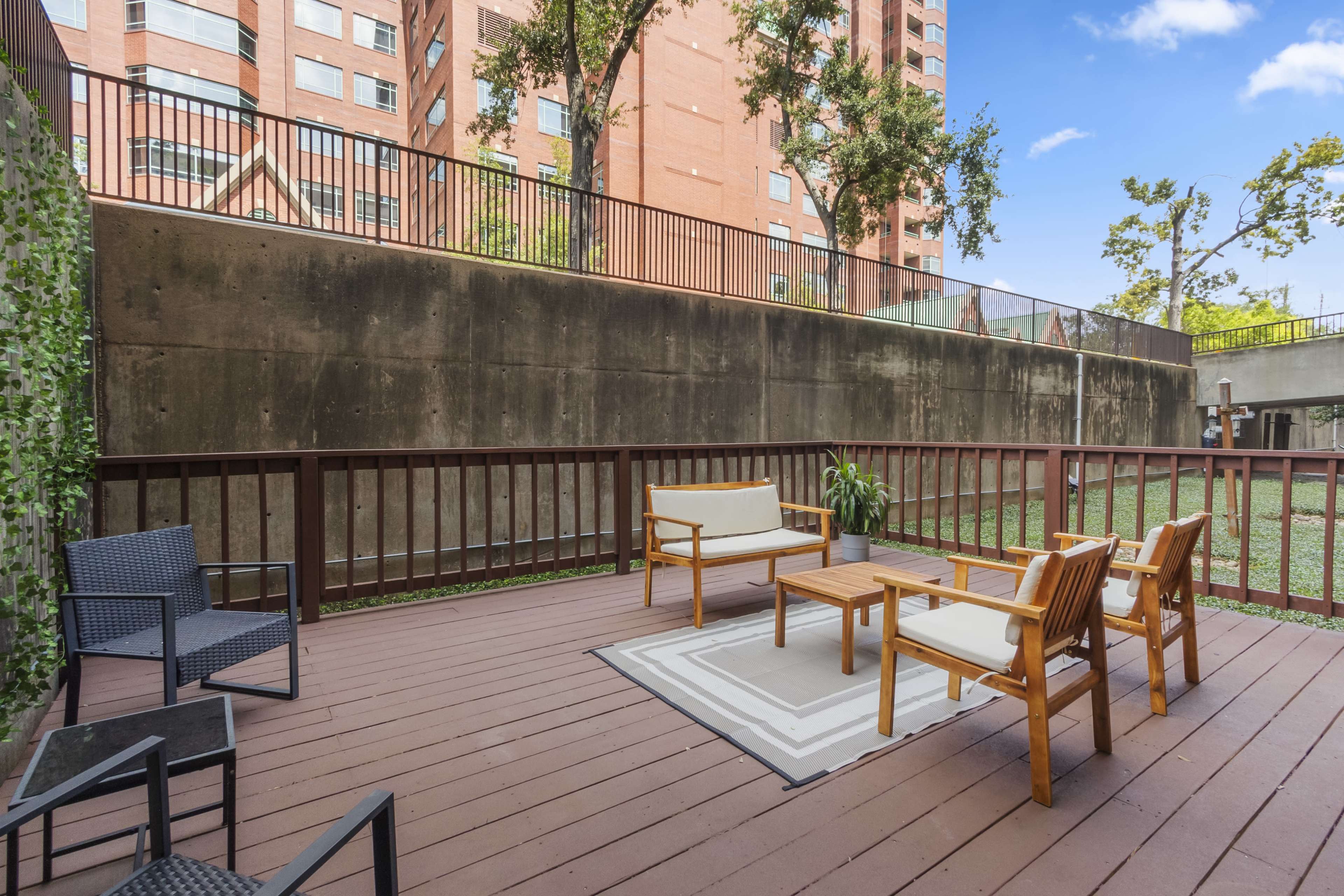 A wooden deck features two chairs, a sofa, and a coffee table, with a backdrop of a tall brick building and greenery along the walls.