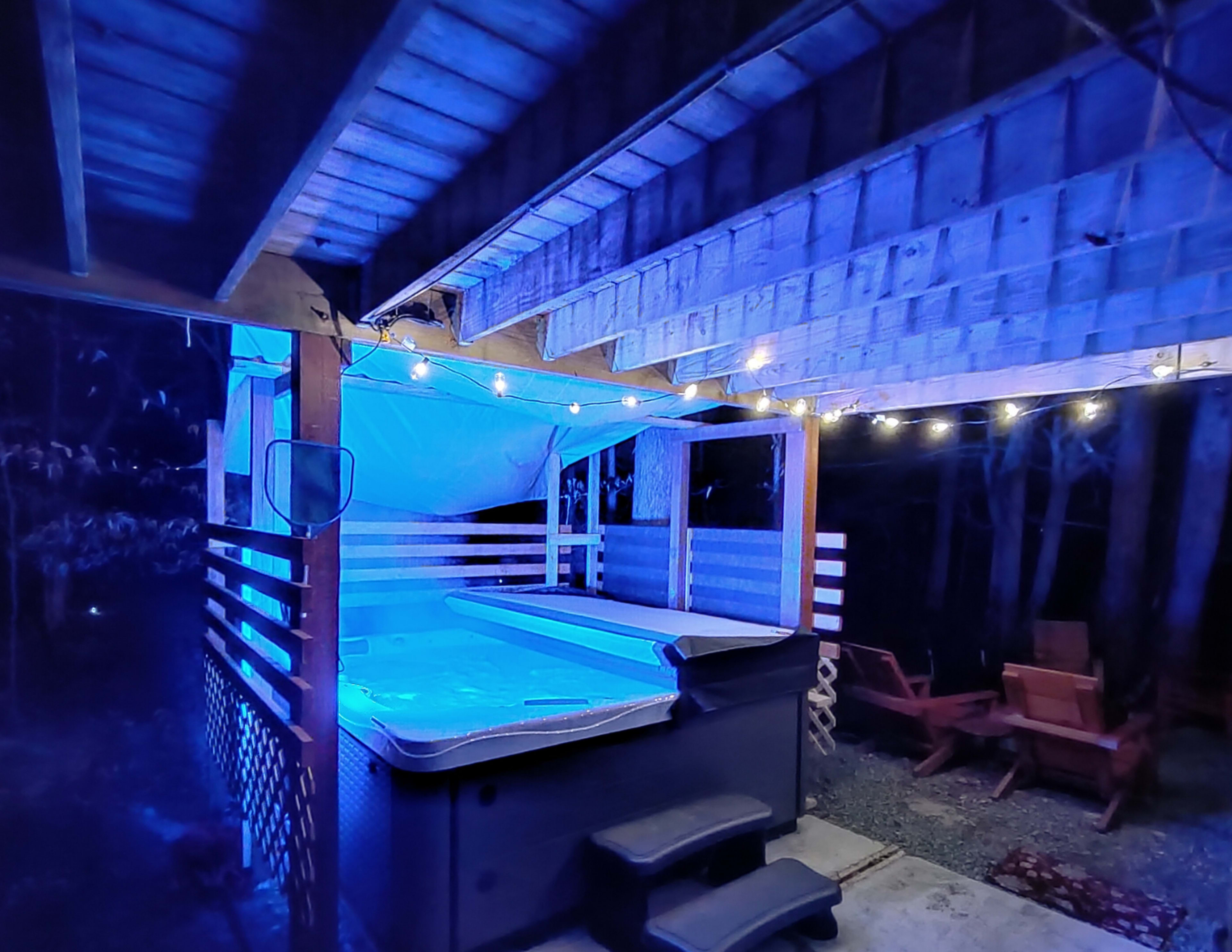 A hot tub is situated under a wooden structure illuminated by blue lights and surrounded by wooden seating and decorative string lights.