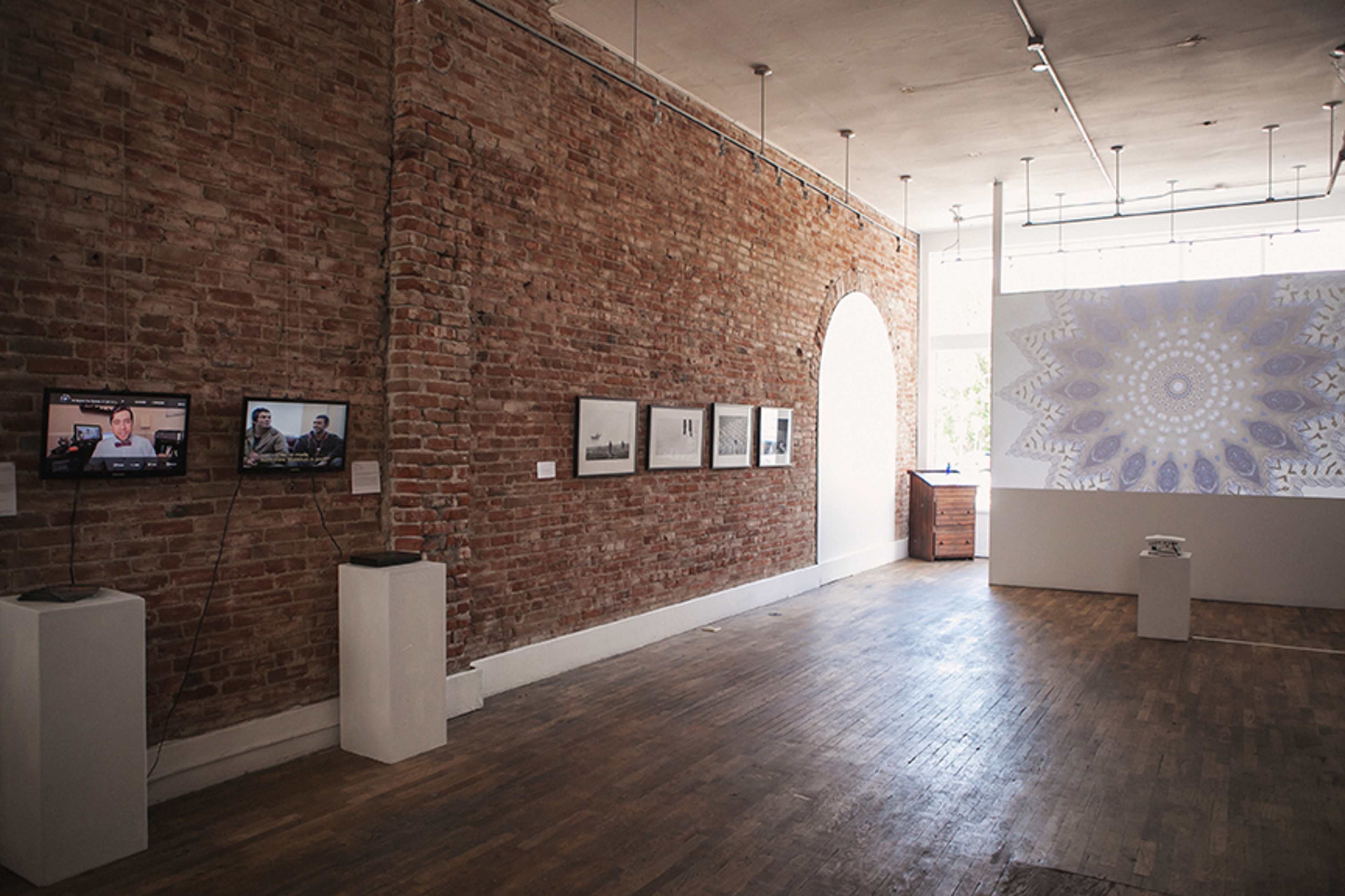 A modern art gallery featuring exposed brick walls, framed photographs, screens displaying video, and a large patterned artwork on the far wall.