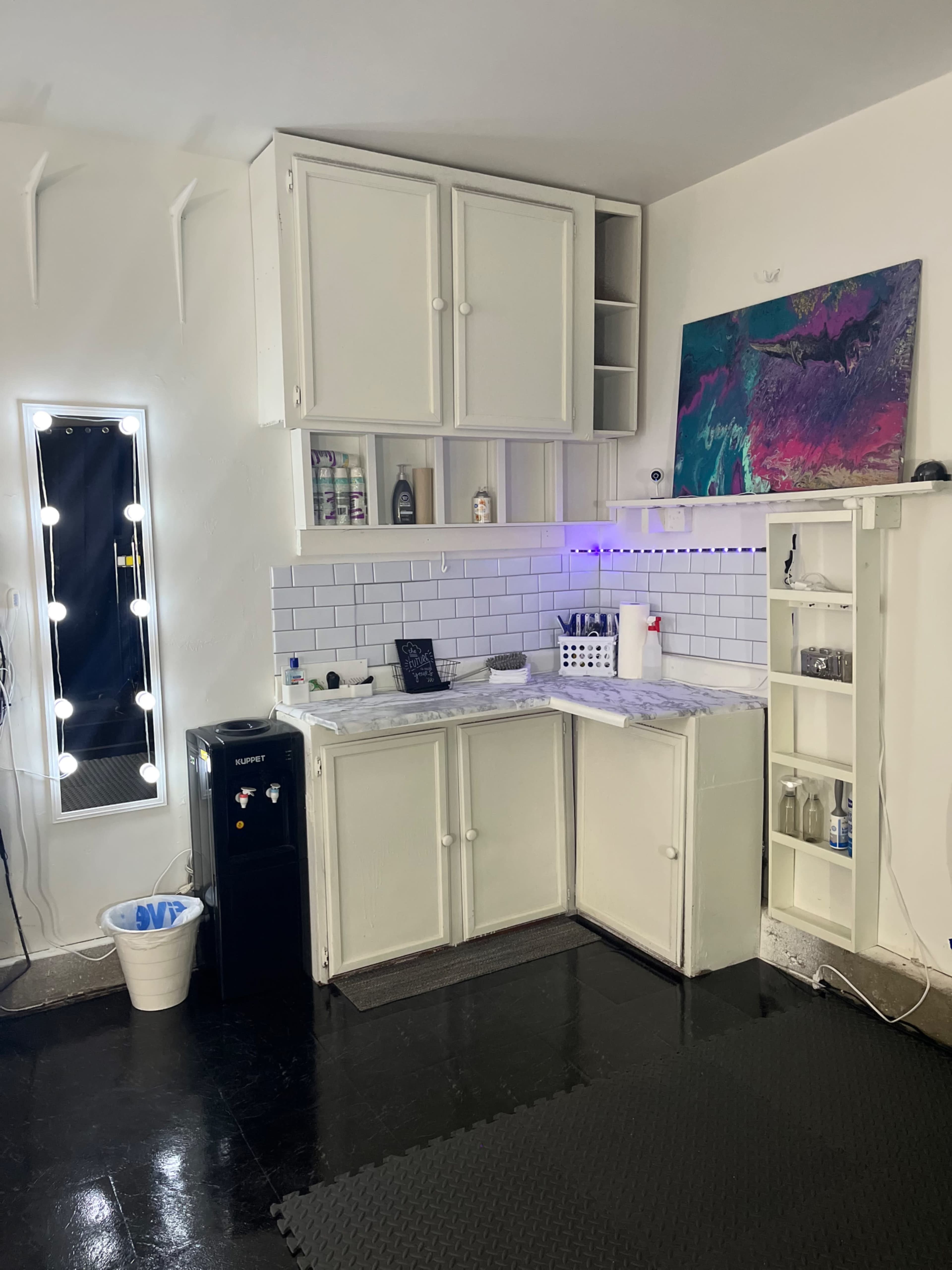 A small, modern salon space featuring a white cabinet setup, a black fridge, and a lit mirror against a stark wall.