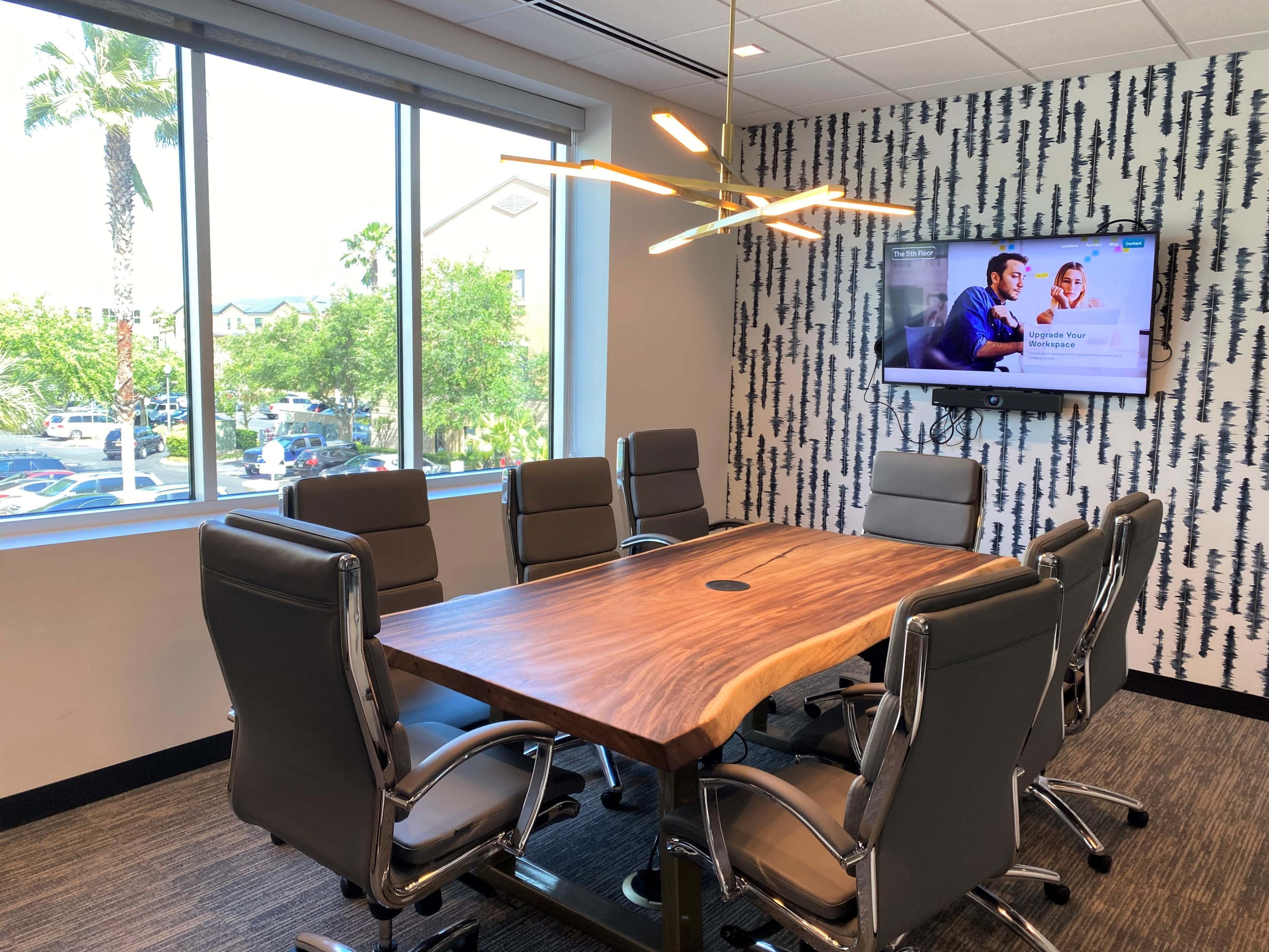 A modern conference room features a large wooden table surrounded by gray leather chairs, with a television displaying content on one wall and large windows offering a view of palm trees outside.