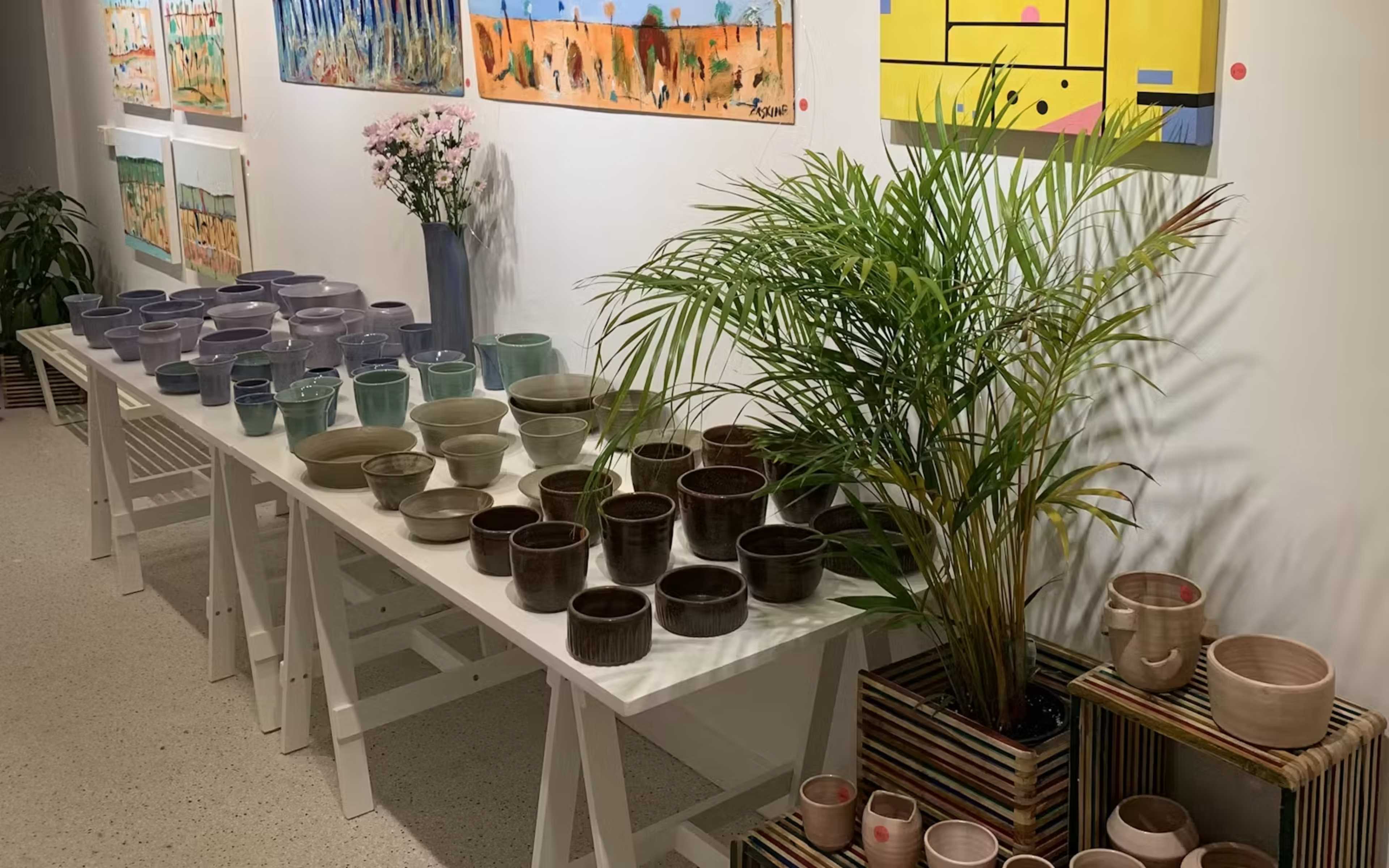A display of various pottery pieces is arranged on tables in an art gallery, accompanied by decorative plants and artwork on the walls.