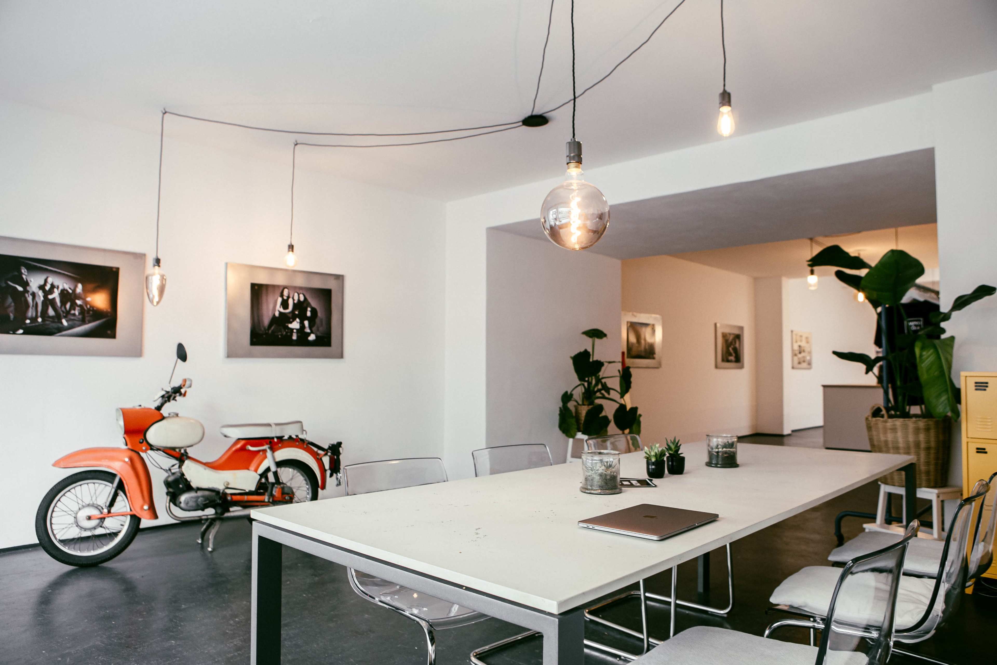 A modern open workspace features a large table surrounded by chairs, with an orange motorcycle displayed against a white wall and plants adding greenery to the space.