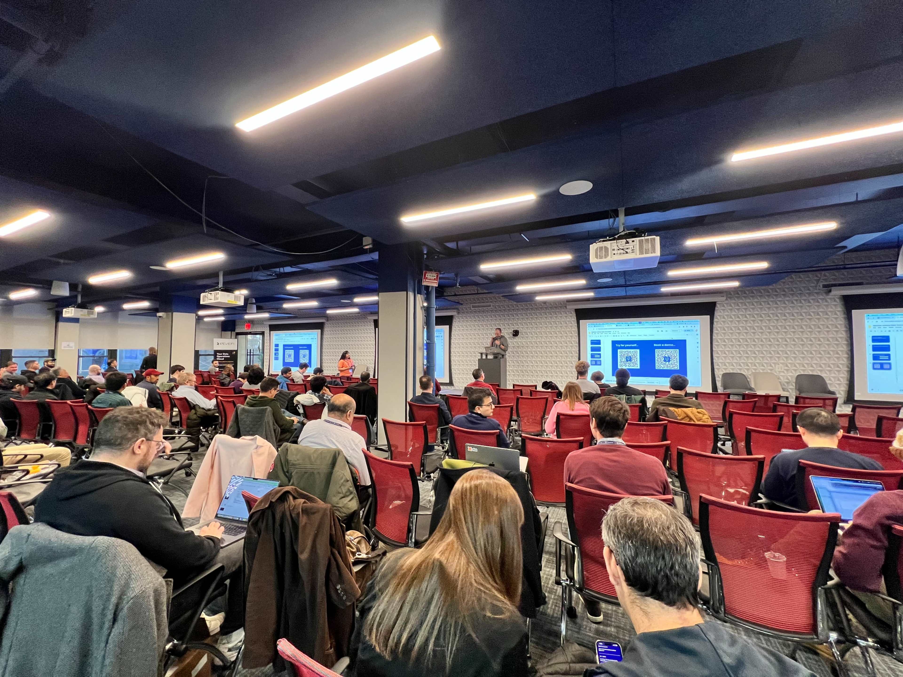 A speaker presents in front of a seated audience in a seminar room equipped with projection screens and laptops.