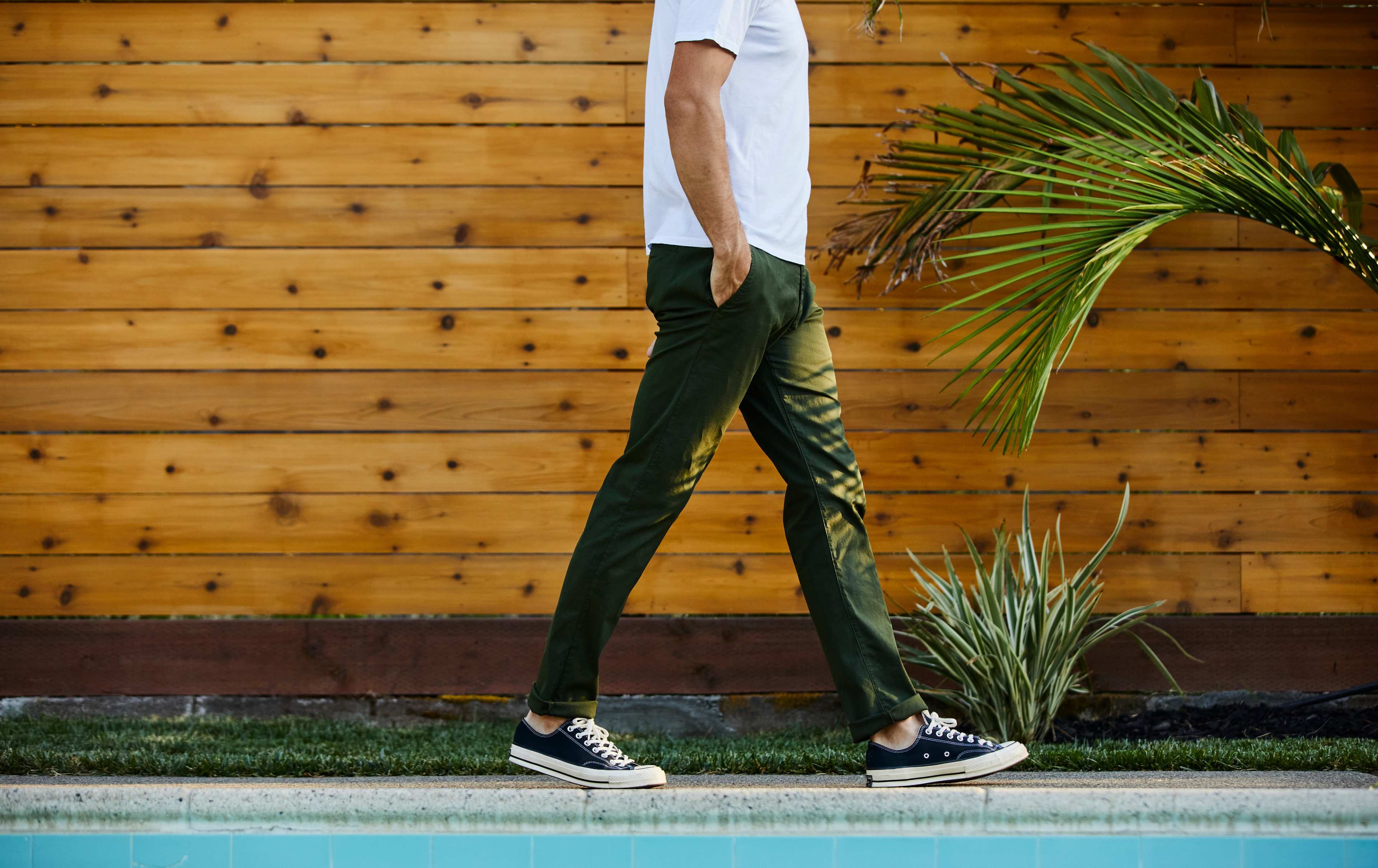 A person wearing a white shirt and green pants walks beside a swimming pool and wooden wall, with plants in the background.