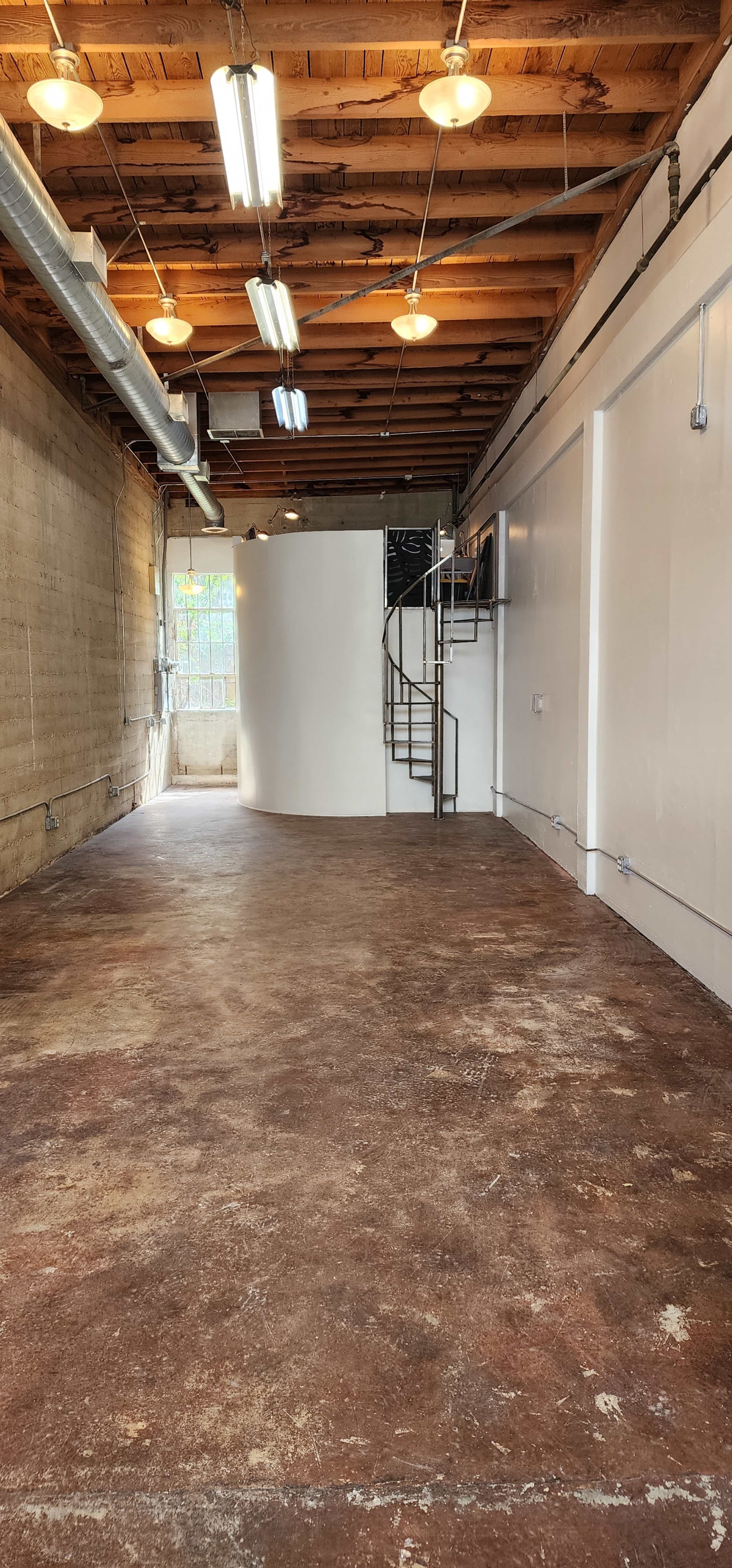 A spacious room with a concrete floor, exposed wooden beams, and a spiral staircase leading to a second level.