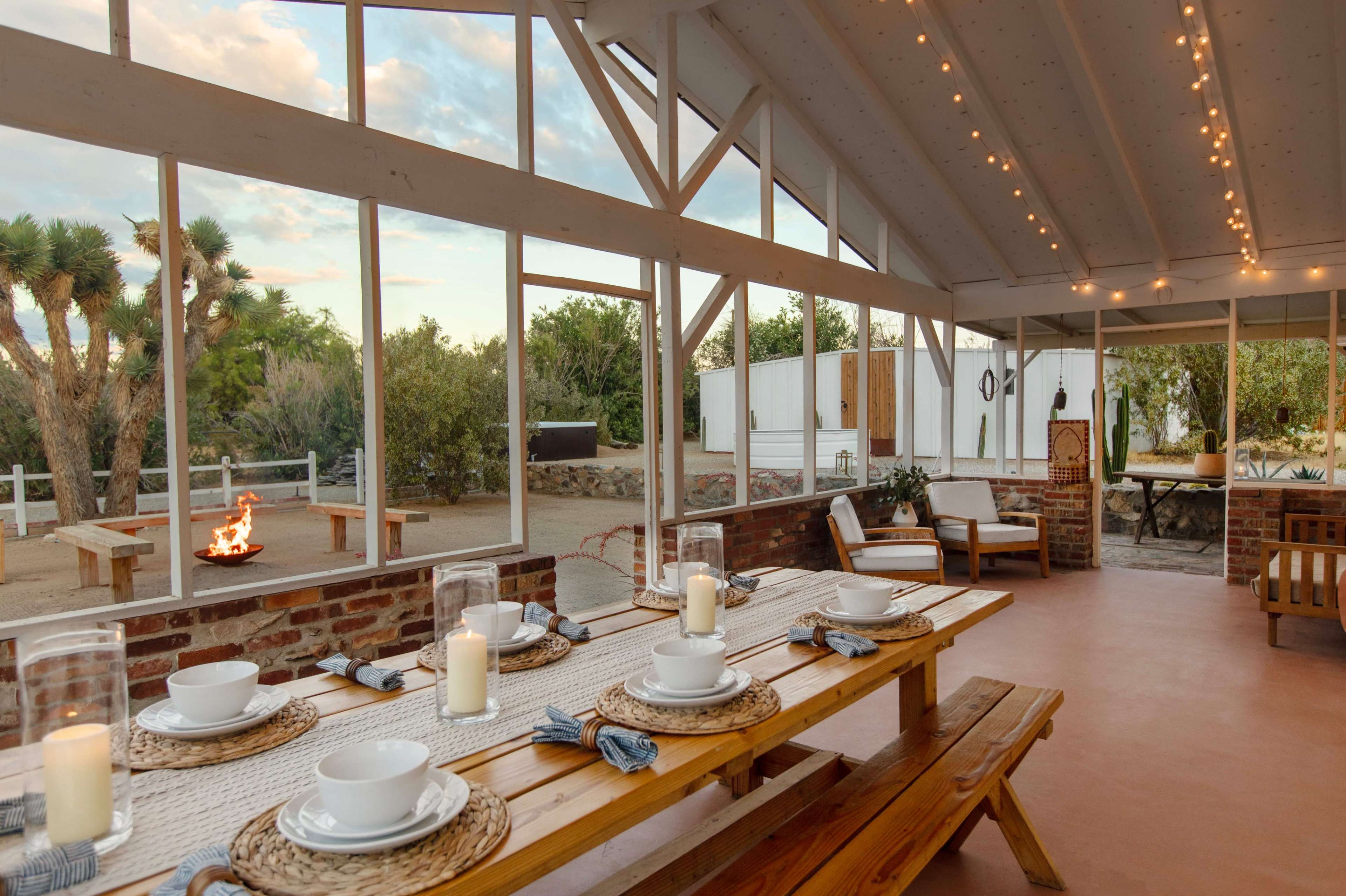 A well-lit dining area with a long wooden table set for a meal, surrounded by large windows, and featuring an outdoor fire pit and lounge area.