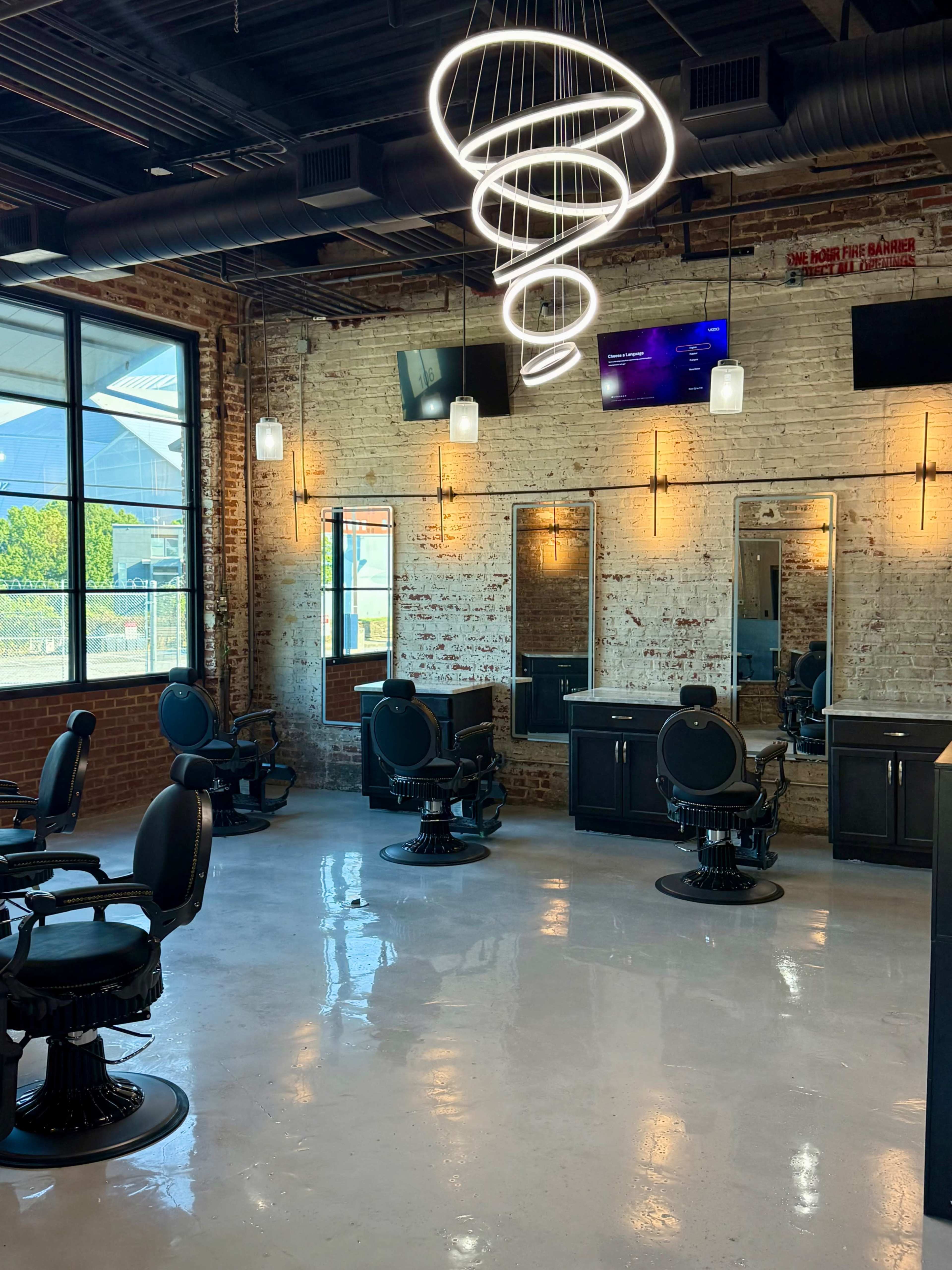 The image shows a modern barbershop with several styling chairs, mirrors, and a minimalist lighting fixture hanging from the ceiling.