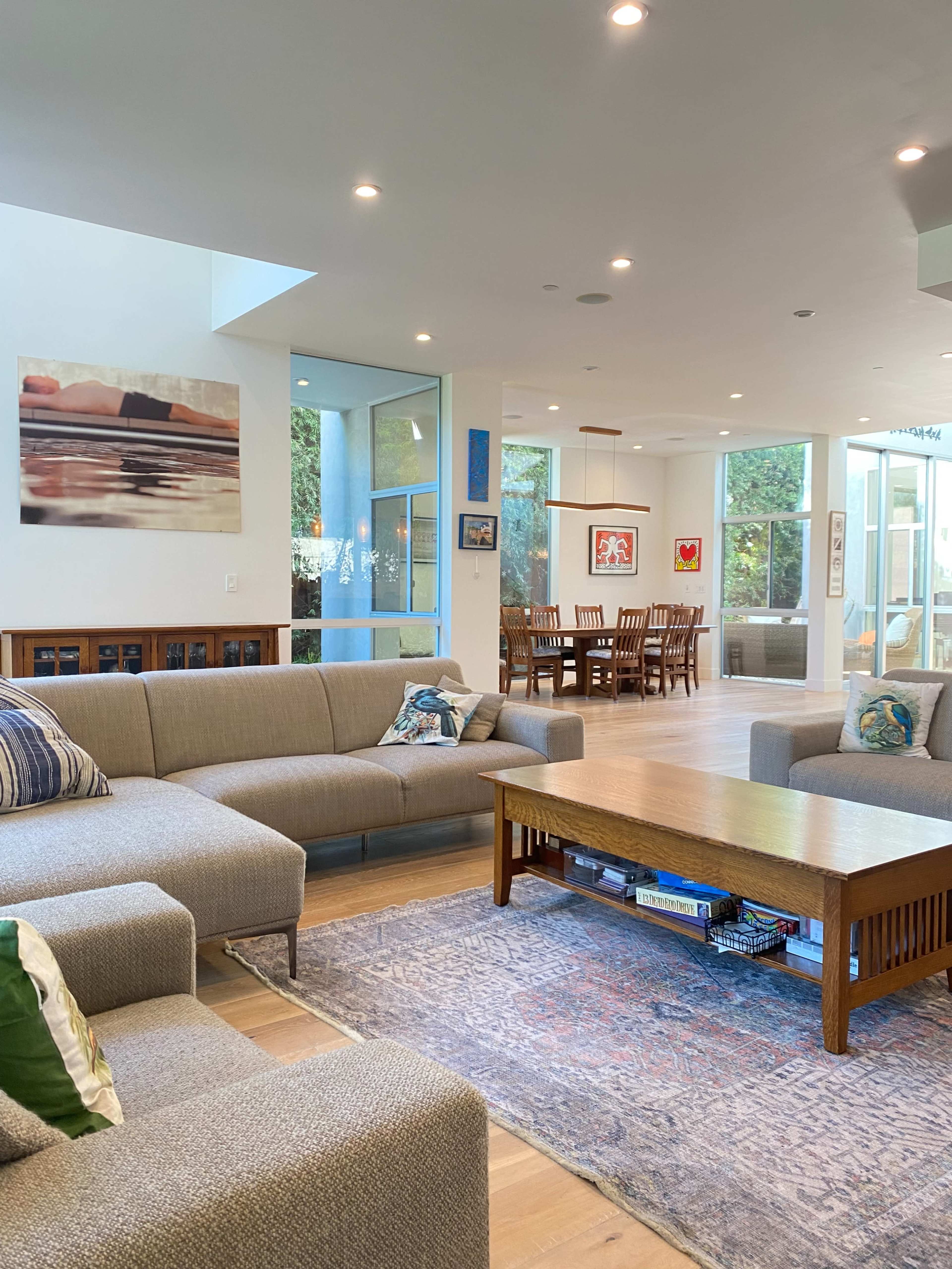 A modern living room with a light gray sectional sofa, a wooden coffee table, and a dining area in the background that includes a wooden dining table and chairs.