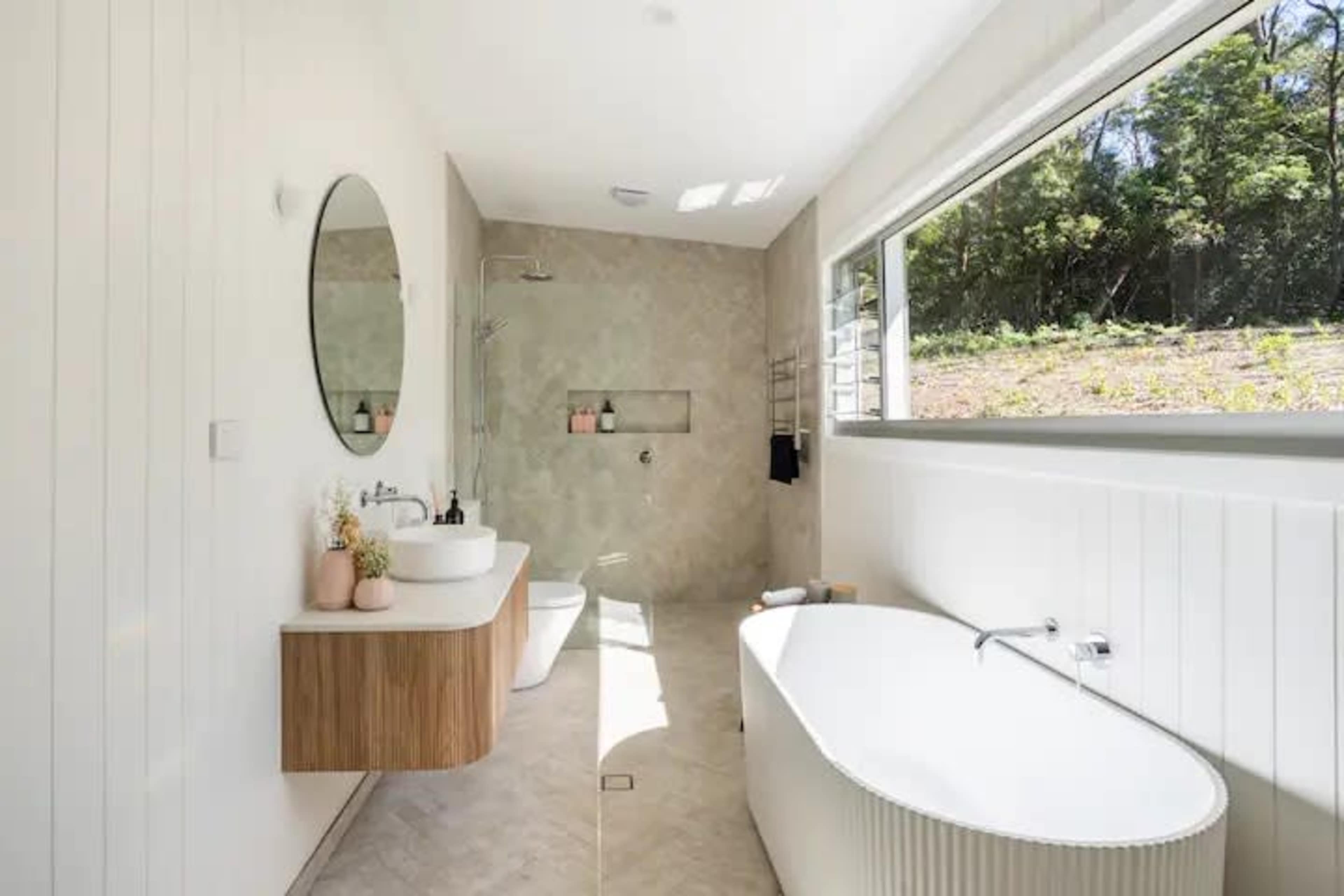 A modern bathroom featuring a freestanding bathtub, a wall-mounted sink with a wooden vanity, and a large window that offers a view of greenery outside.