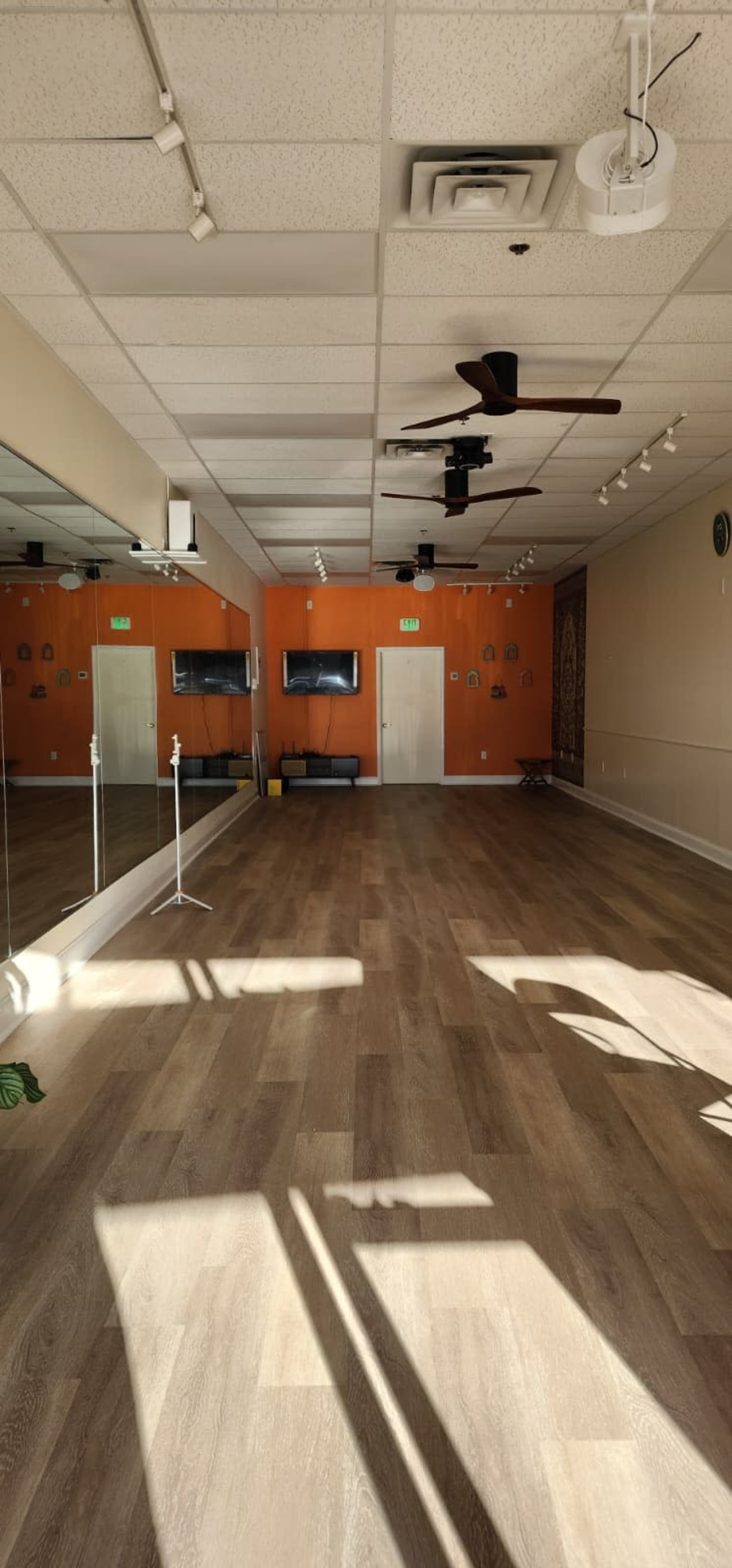 The image shows an empty studio space with wooden flooring, mirrors on one wall, and orange accent walls, featuring two televisions and a ceiling fan.