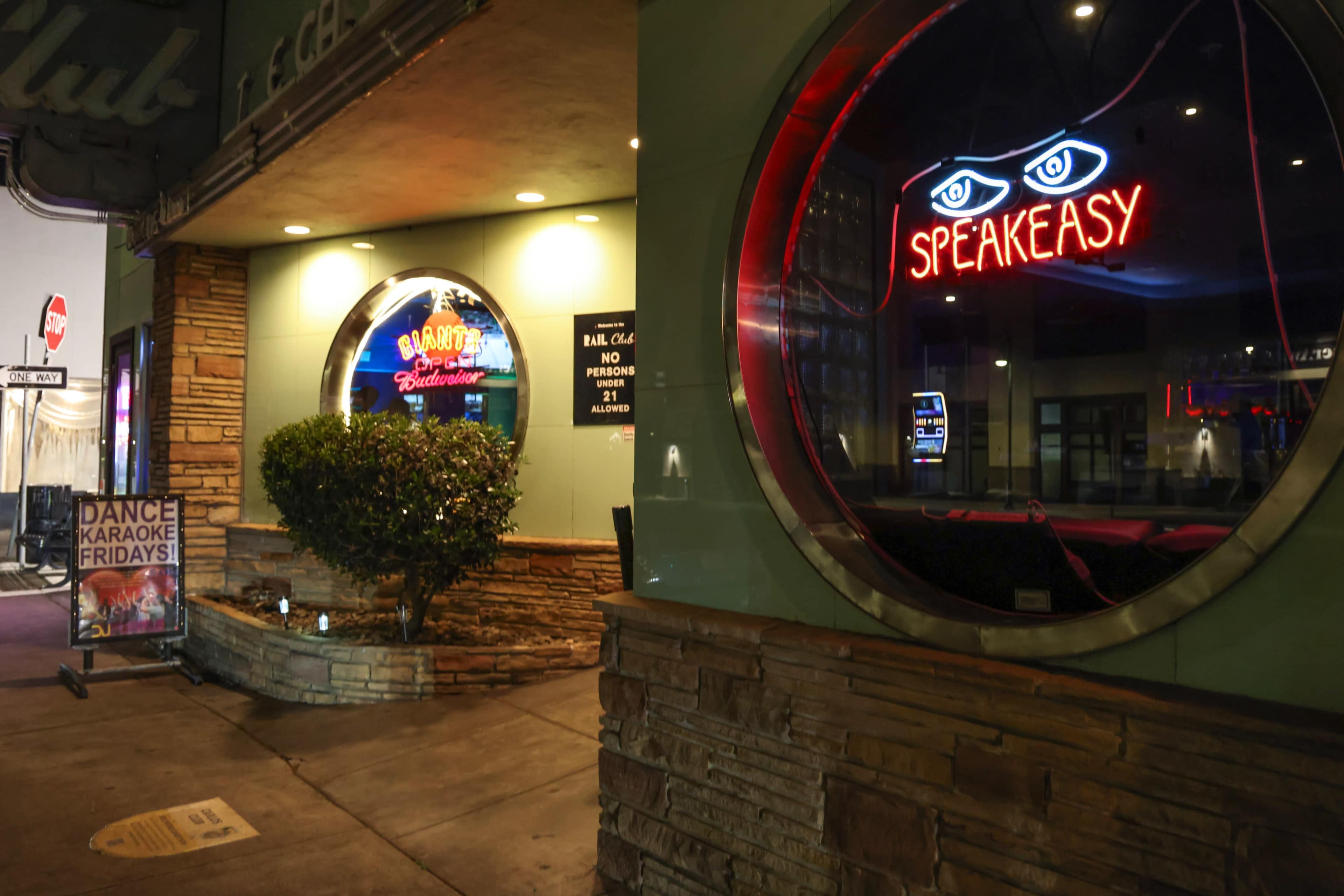 The image shows a brightly lit entrance of a club featuring a neon "SPEAKEASY" sign and a small bush outside.