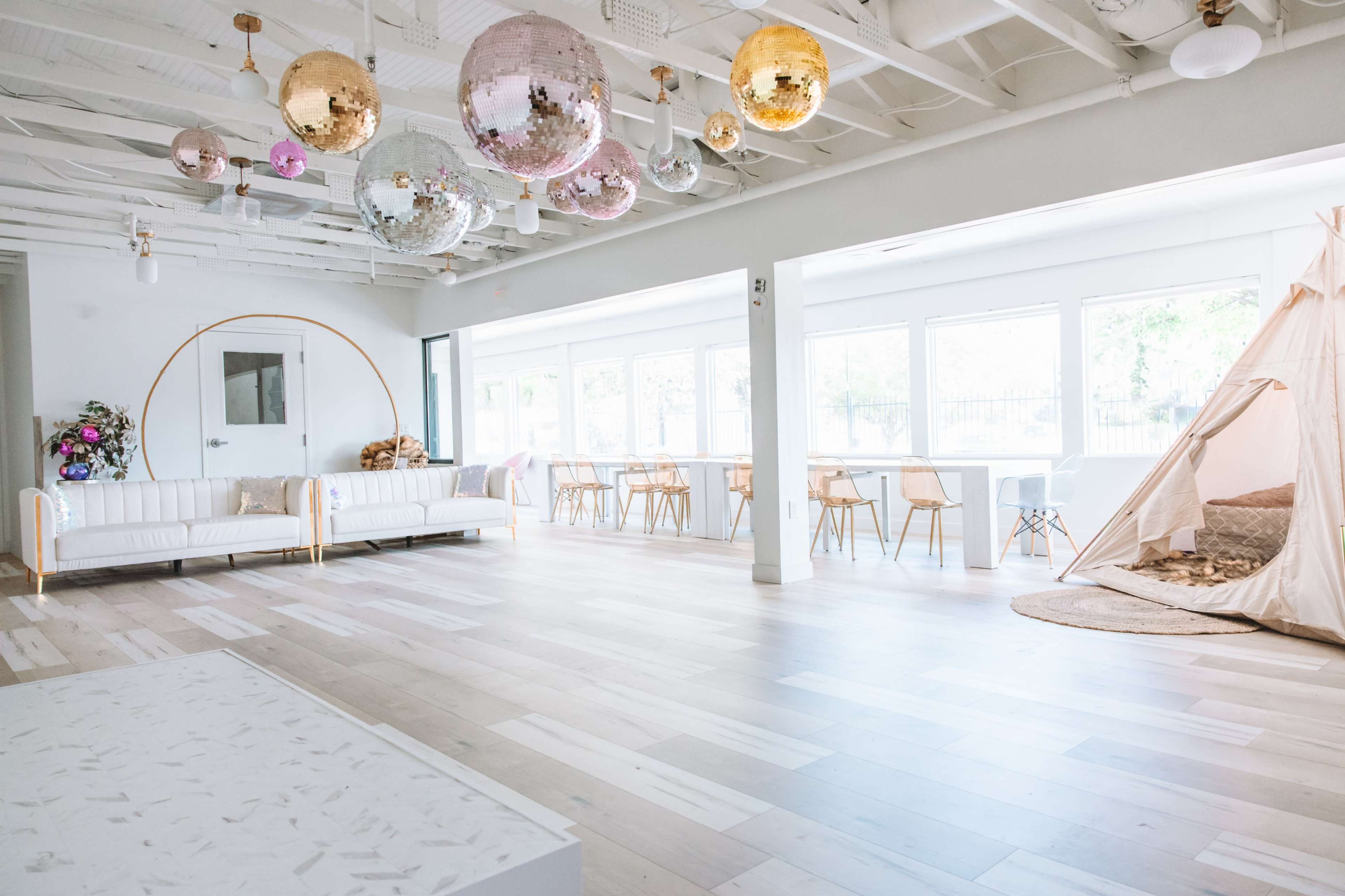 A bright, spacious room with wooden floors, decorated with hanging disco balls and a cozy teepee, featuring white sofas and wooden dining tables.