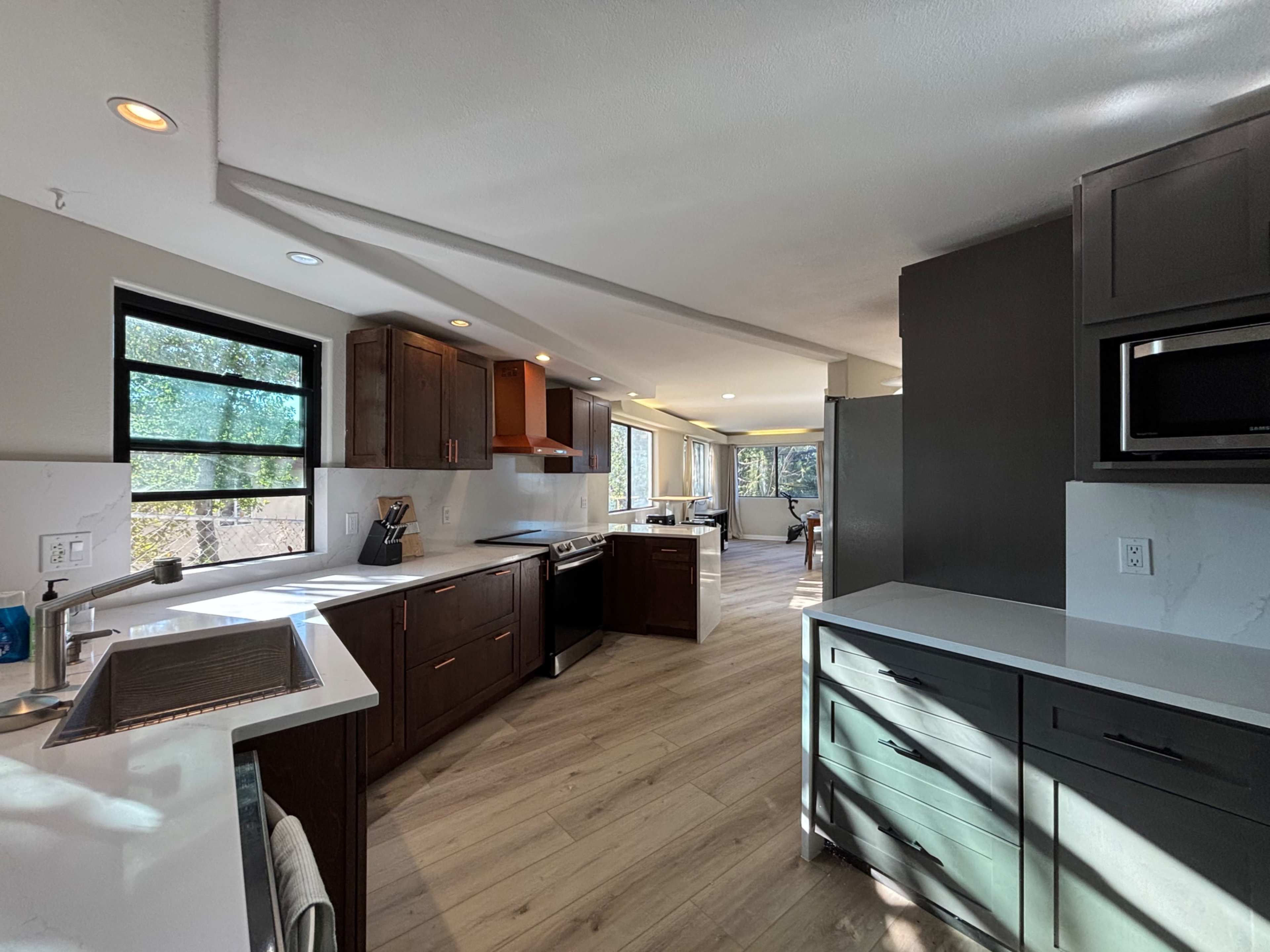 The image shows a modern kitchen with dark wood cabinets, white countertops, and large windows allowing natural light to enter.
