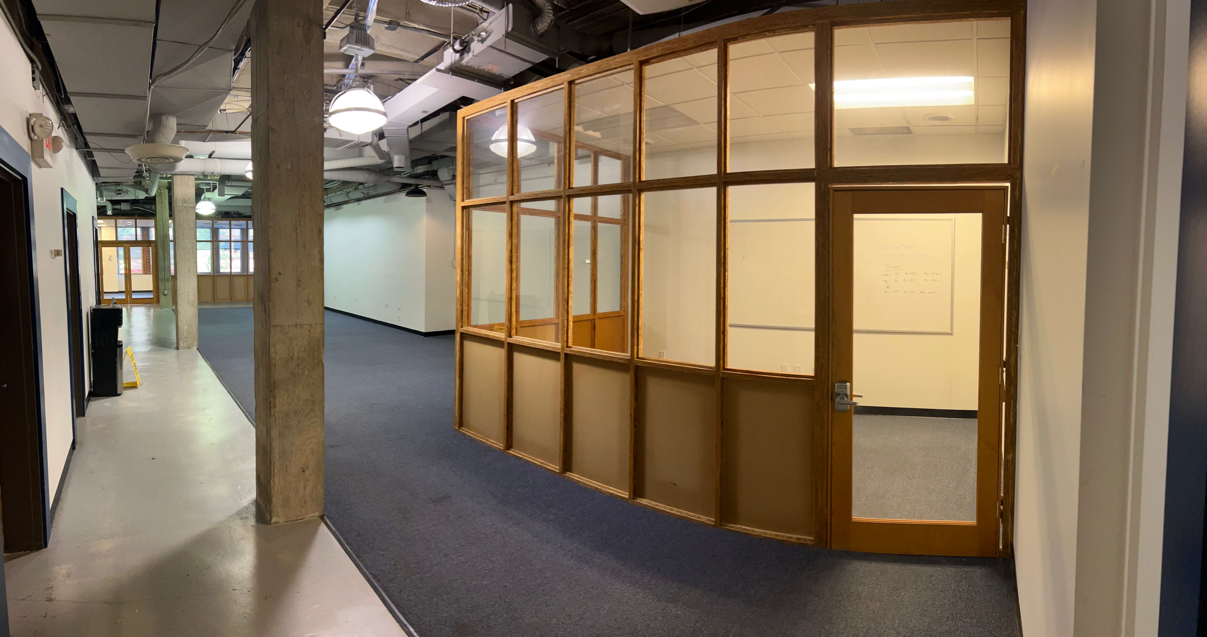 A hallway with a glass-paned room on the right and a door leading to the room, surrounded by bare walls and a carpeted floor.