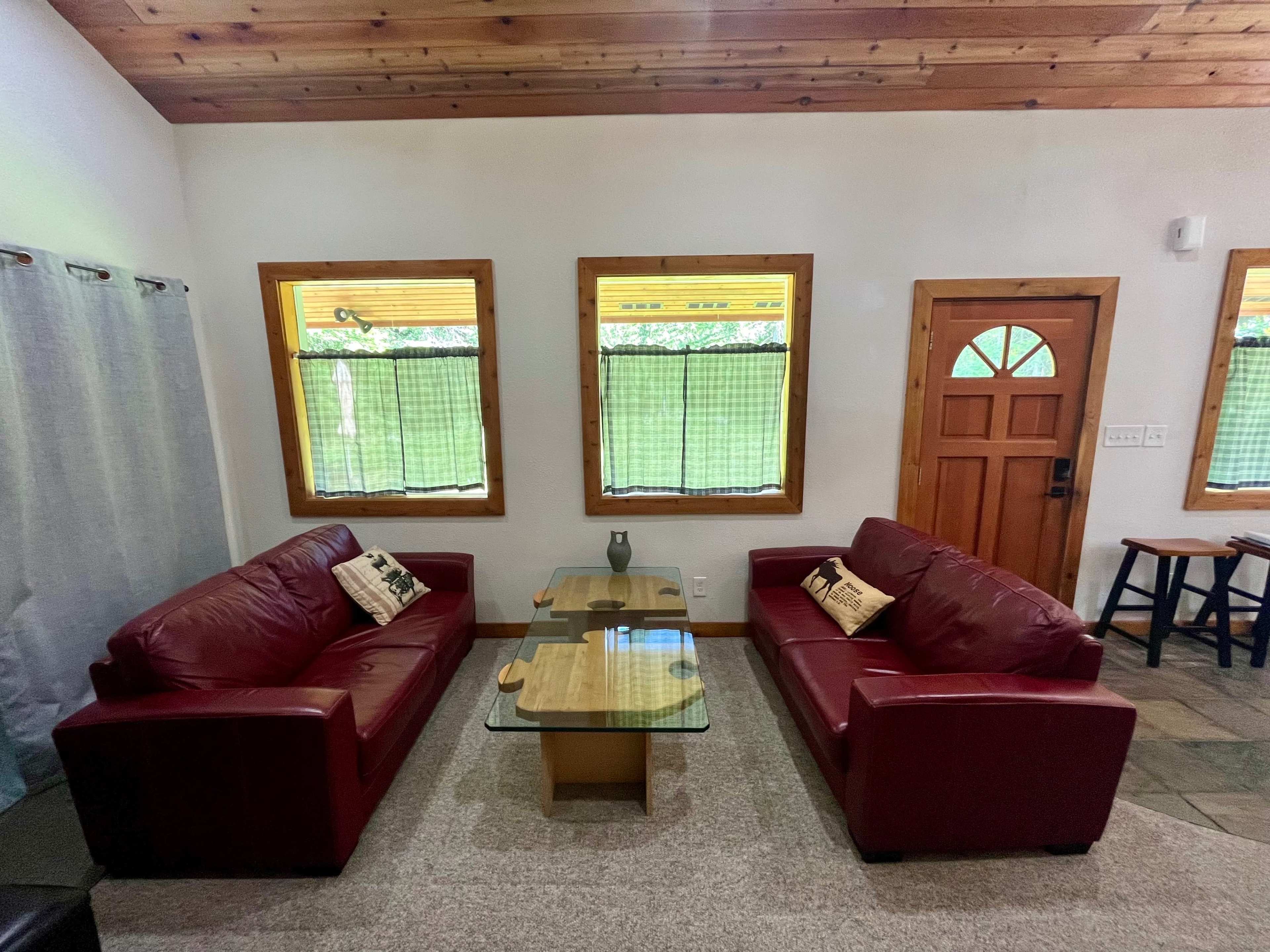 A living room with two red sofas facing a glass coffee table, framed by windows with green curtains and a wooden door.