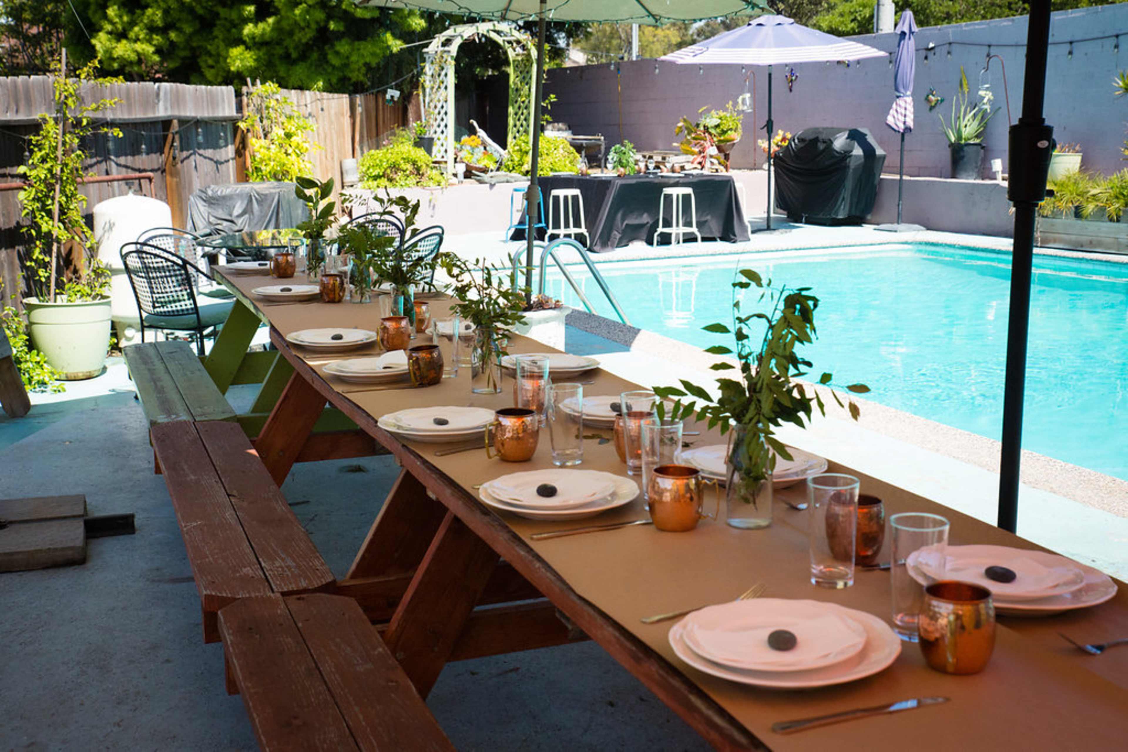 A long wooden table is set with dishes and glasses beside a swimming pool in a backyard garden.
