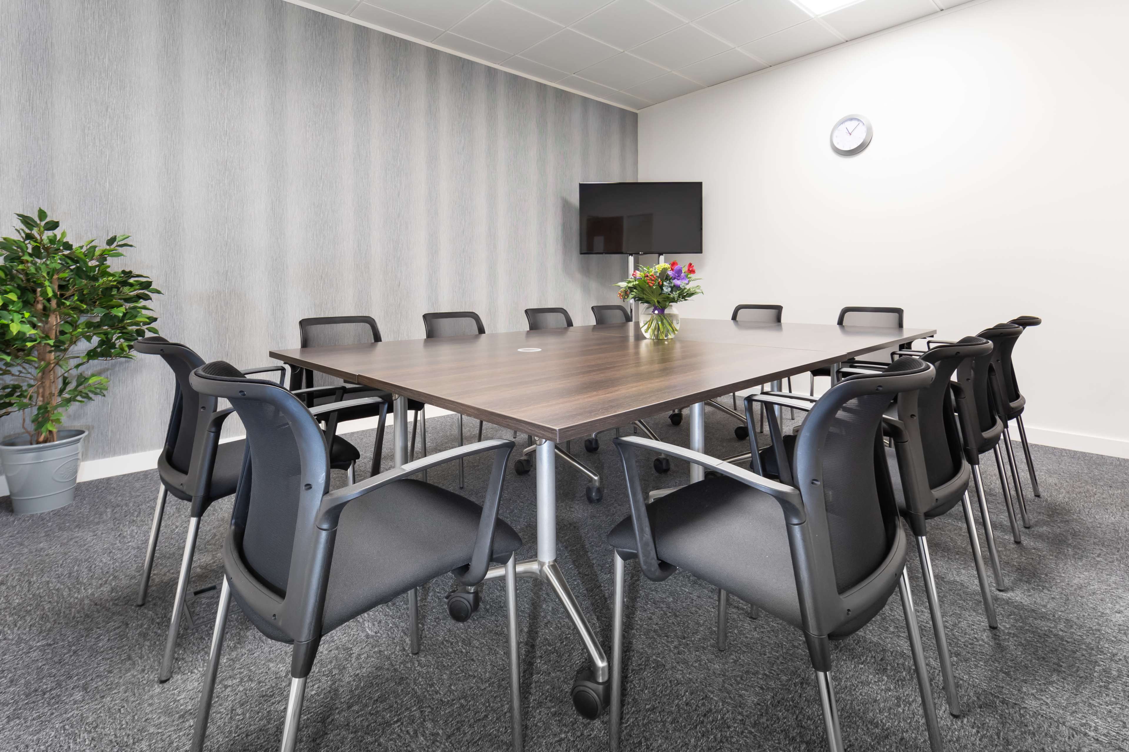 A conference room features a large wooden table surrounded by black chairs, with a television mounted on the wall and a clock above.