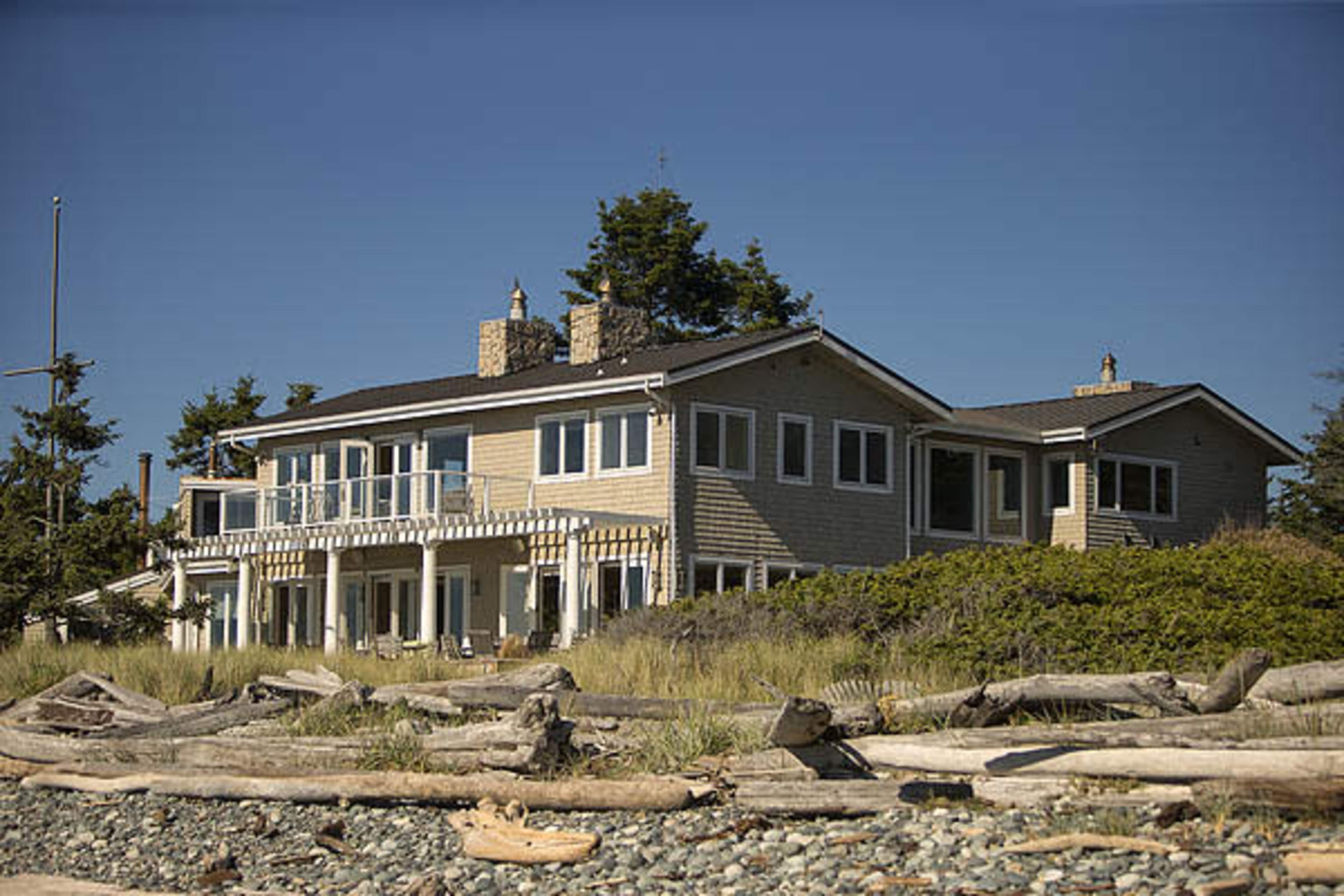 A large beach house with multiple levels and a wooden deck sits near the shore, surrounded by sand and driftwood.