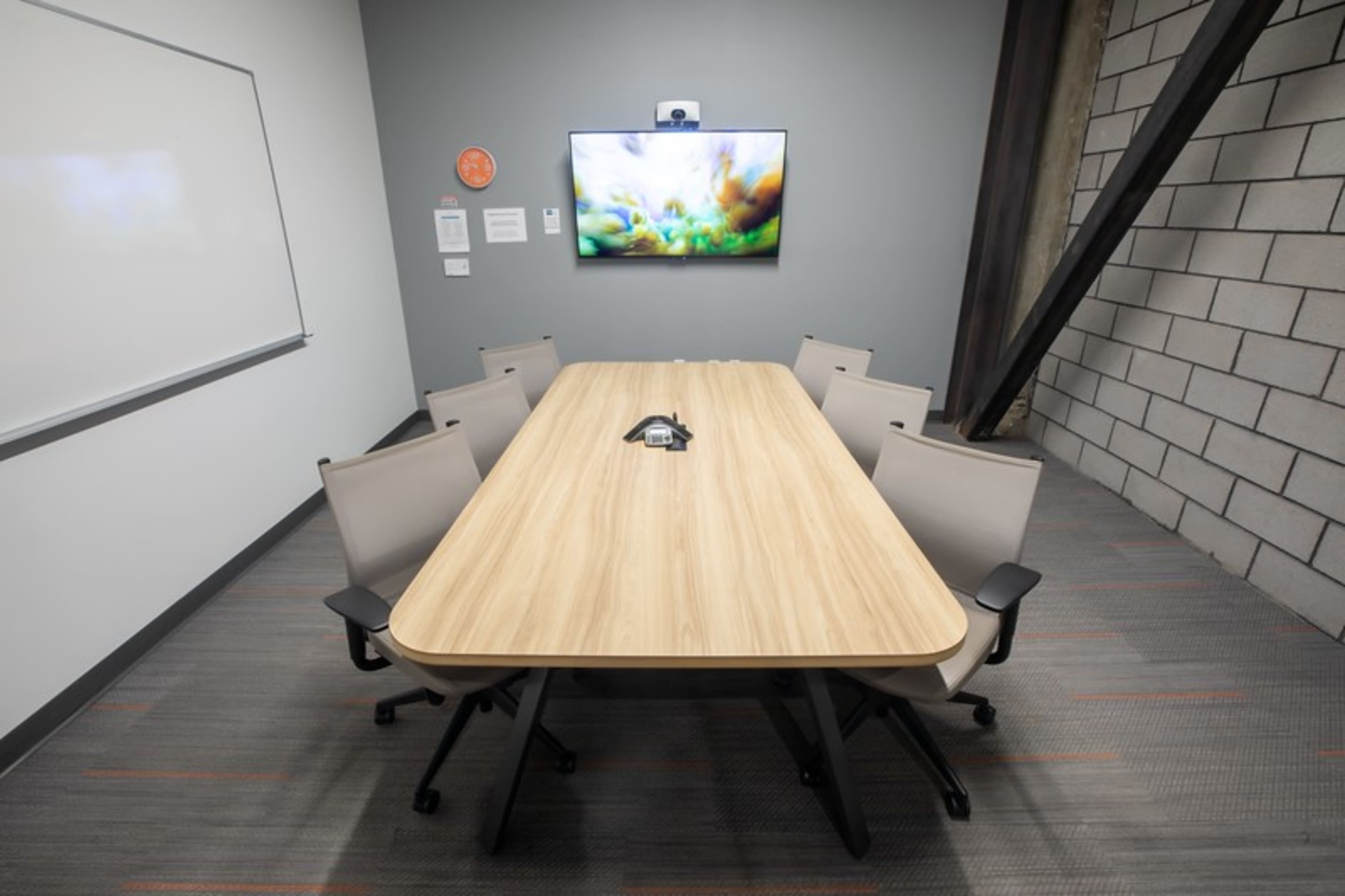 A conference room features a long wooden table surrounded by six chairs, with a wall-mounted screen displaying a colorful image.