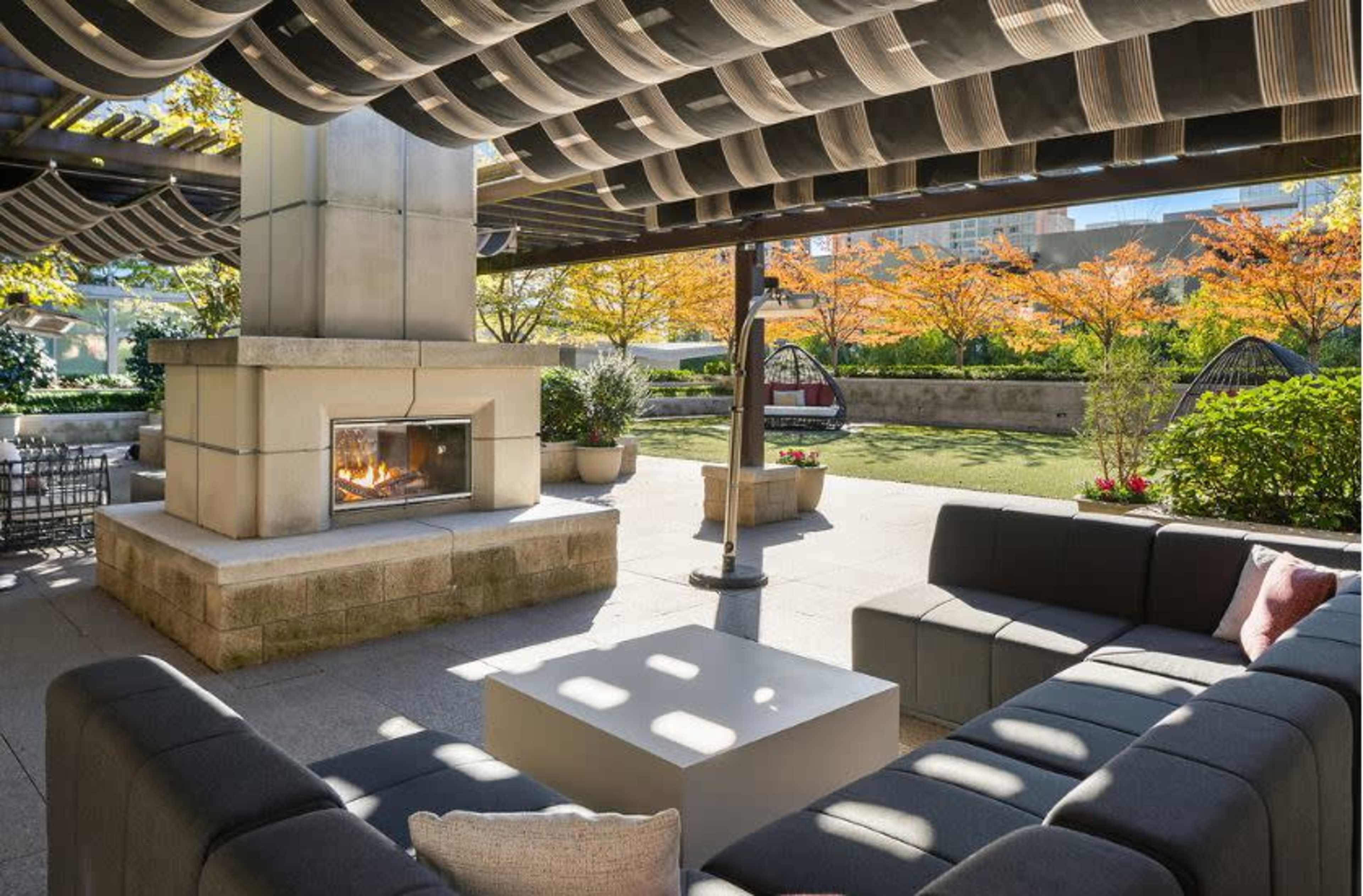 A modern outdoor seating area with a fireplace, featuring sectional sofas and decorative plants, under a shaded pergola.