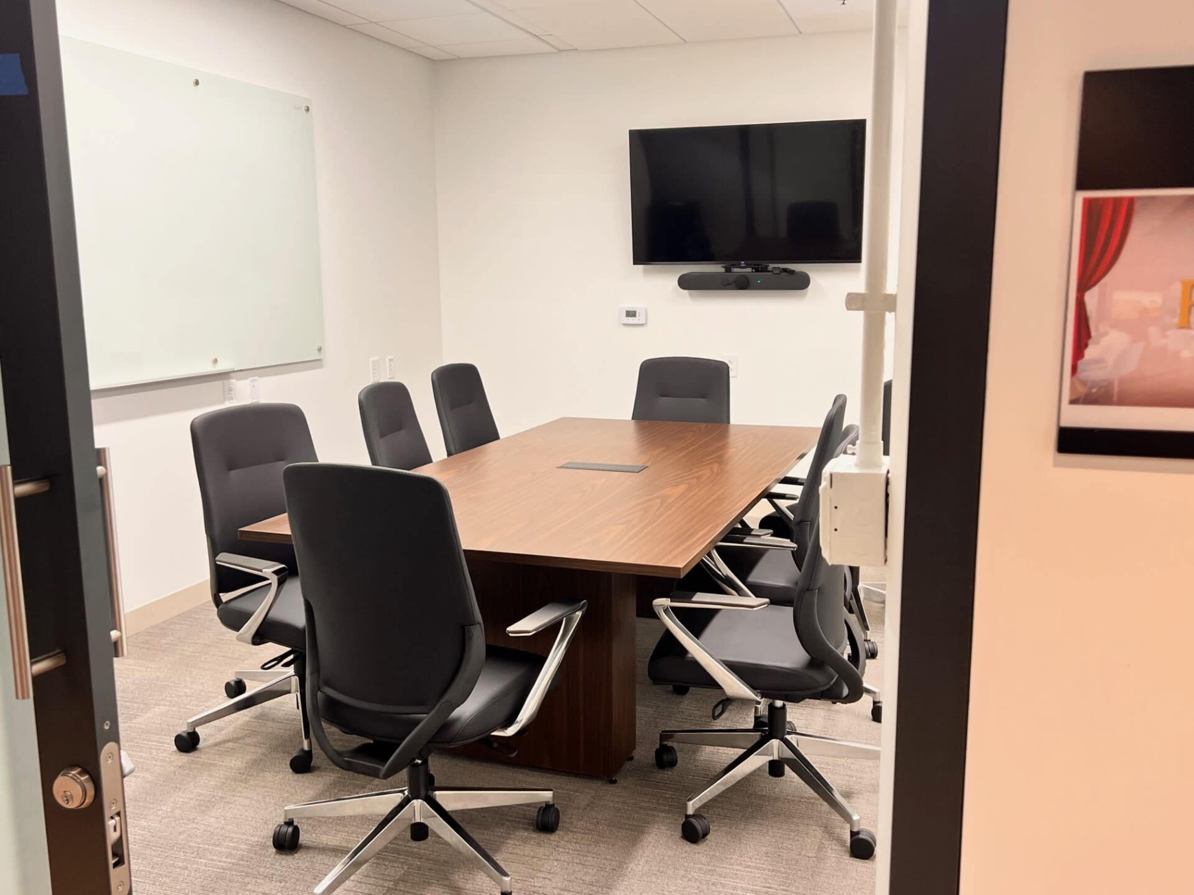 A conference room features a large wooden table surrounded by six ergonomic chairs, with a whiteboard and a television on the walls.