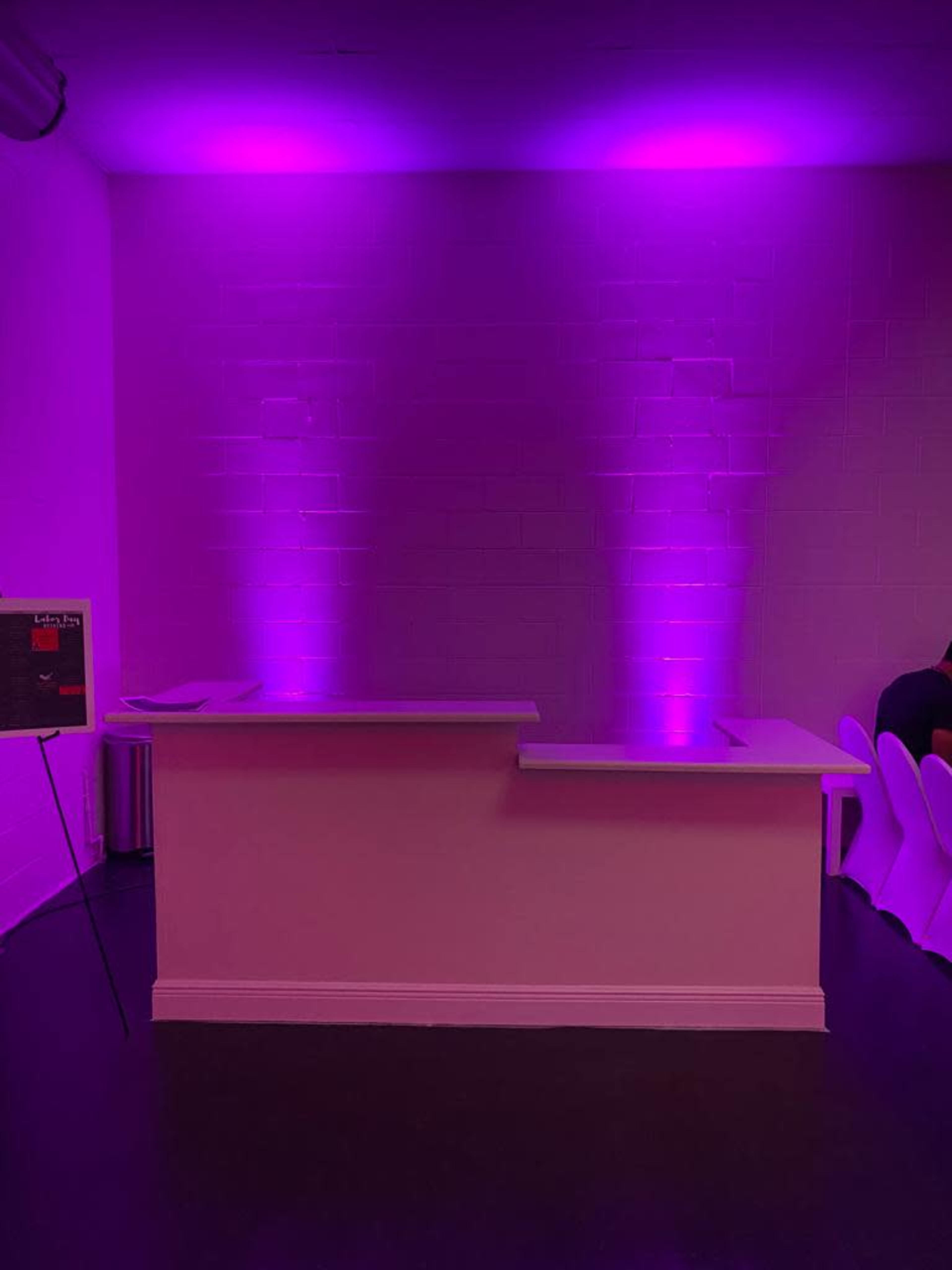 A simple bar area with a white counter is illuminated by two purple lights in a dimly lit room.