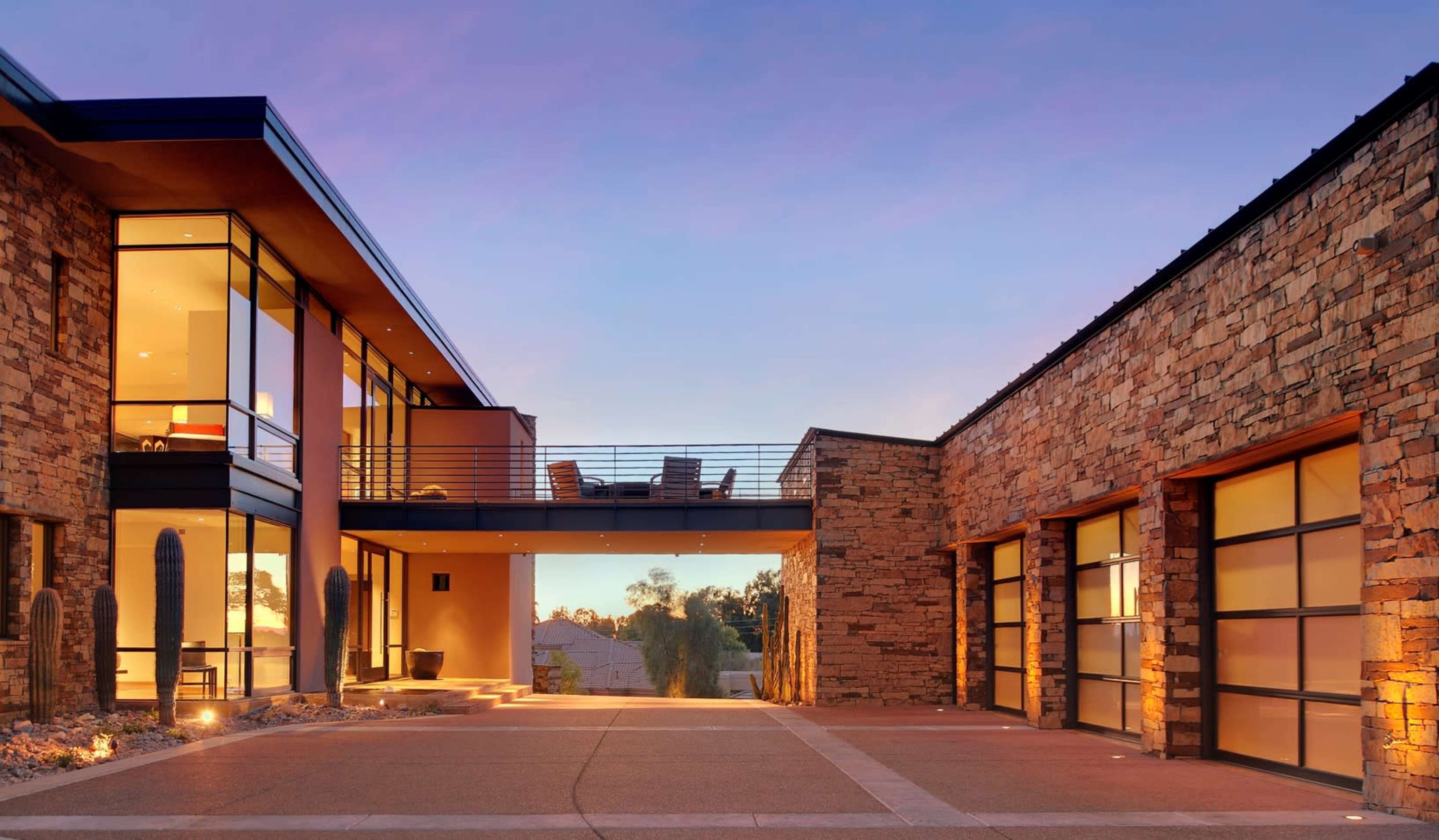 A modern stone and glass house features a bridge connecting two sections, with a driveway leading up to it at dusk.