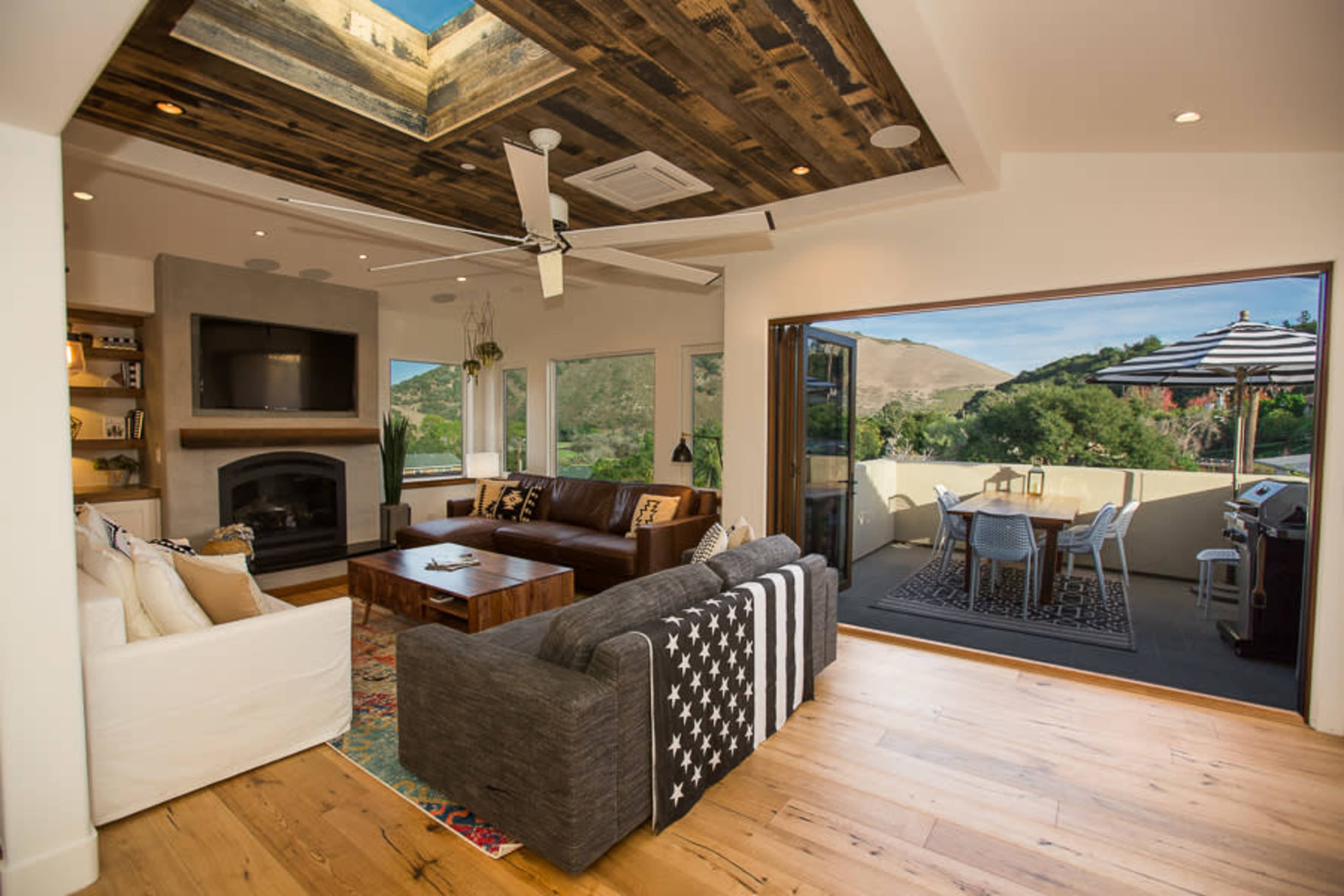 A modern living room with a seating area, a television above a fireplace, and large doors leading to a patio with outdoor dining furniture.