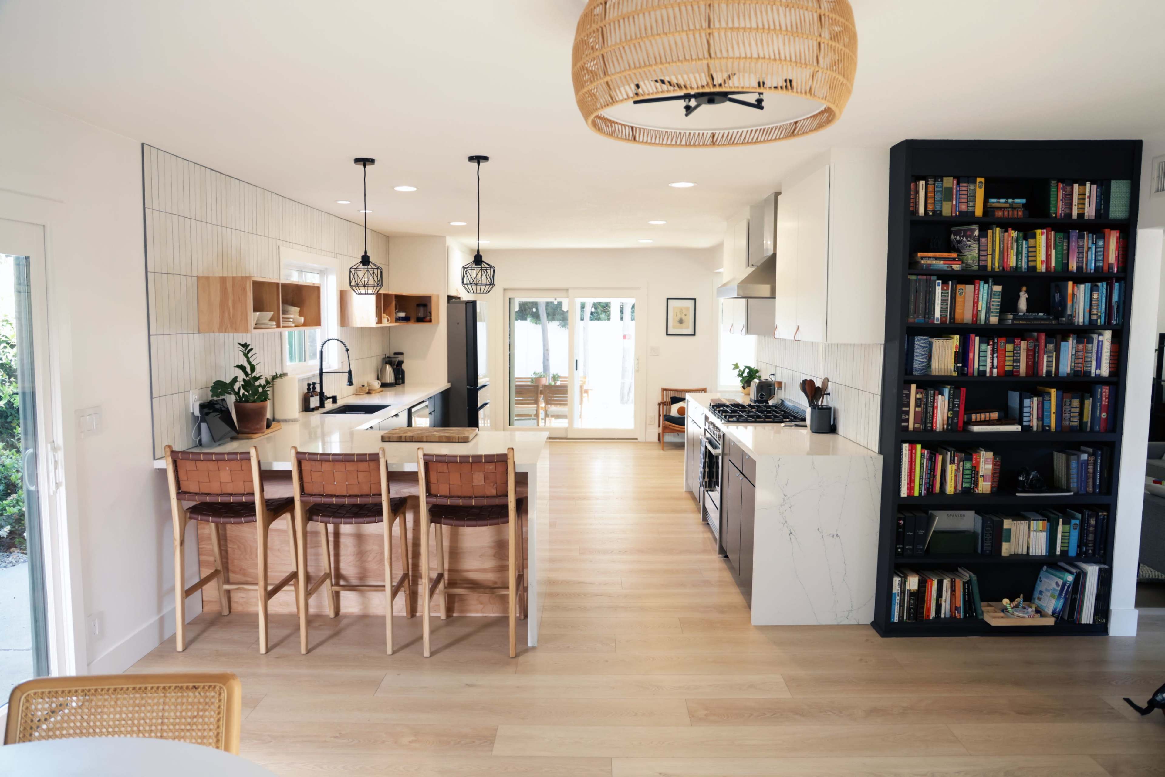 A modern kitchen with wooden flooring, a breakfast bar with wooden stools, and a bookshelf filled with books.