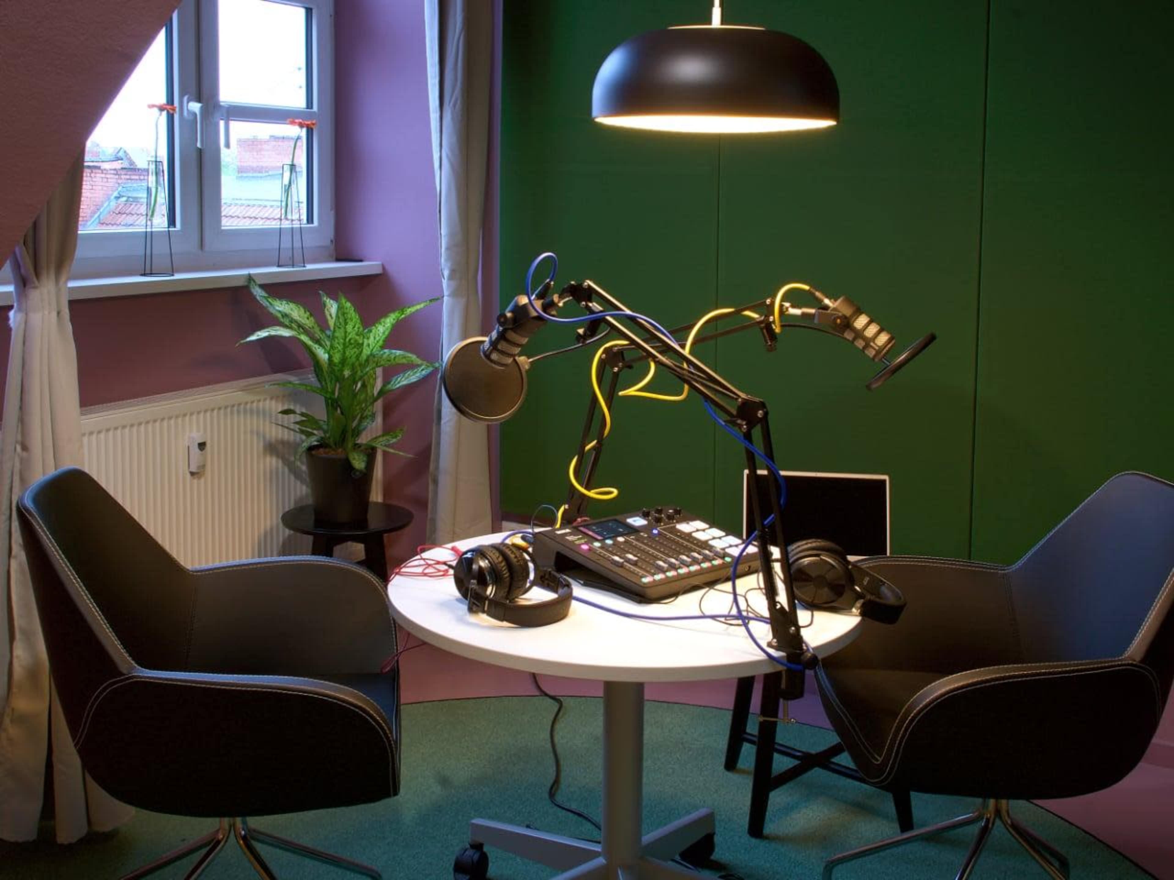 A small recording studio is set up with two chairs, a table containing audio equipment, and microphones, all under a hanging light against a green background.