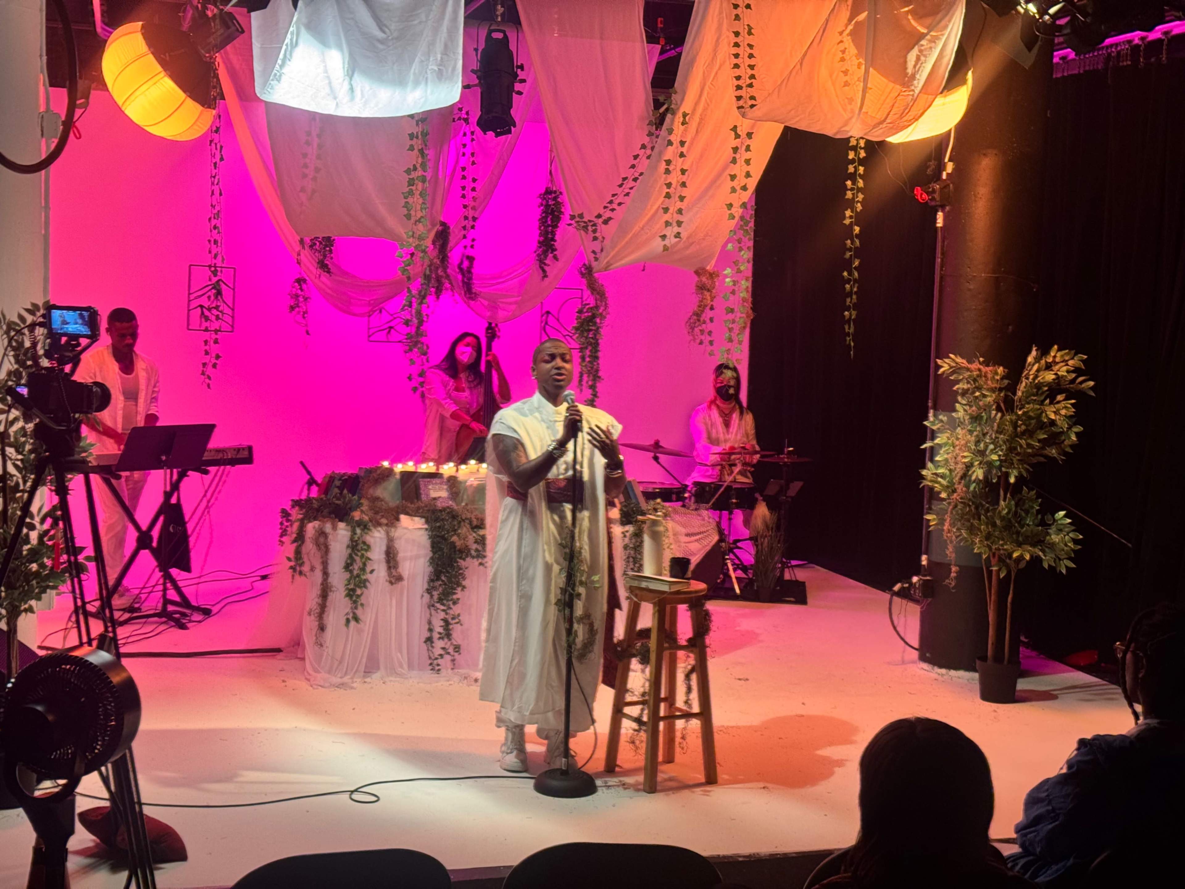 A performer stands on a stool in a brightly lit venue adorned with draped fabric and greenery while musicians play instruments in the background.
