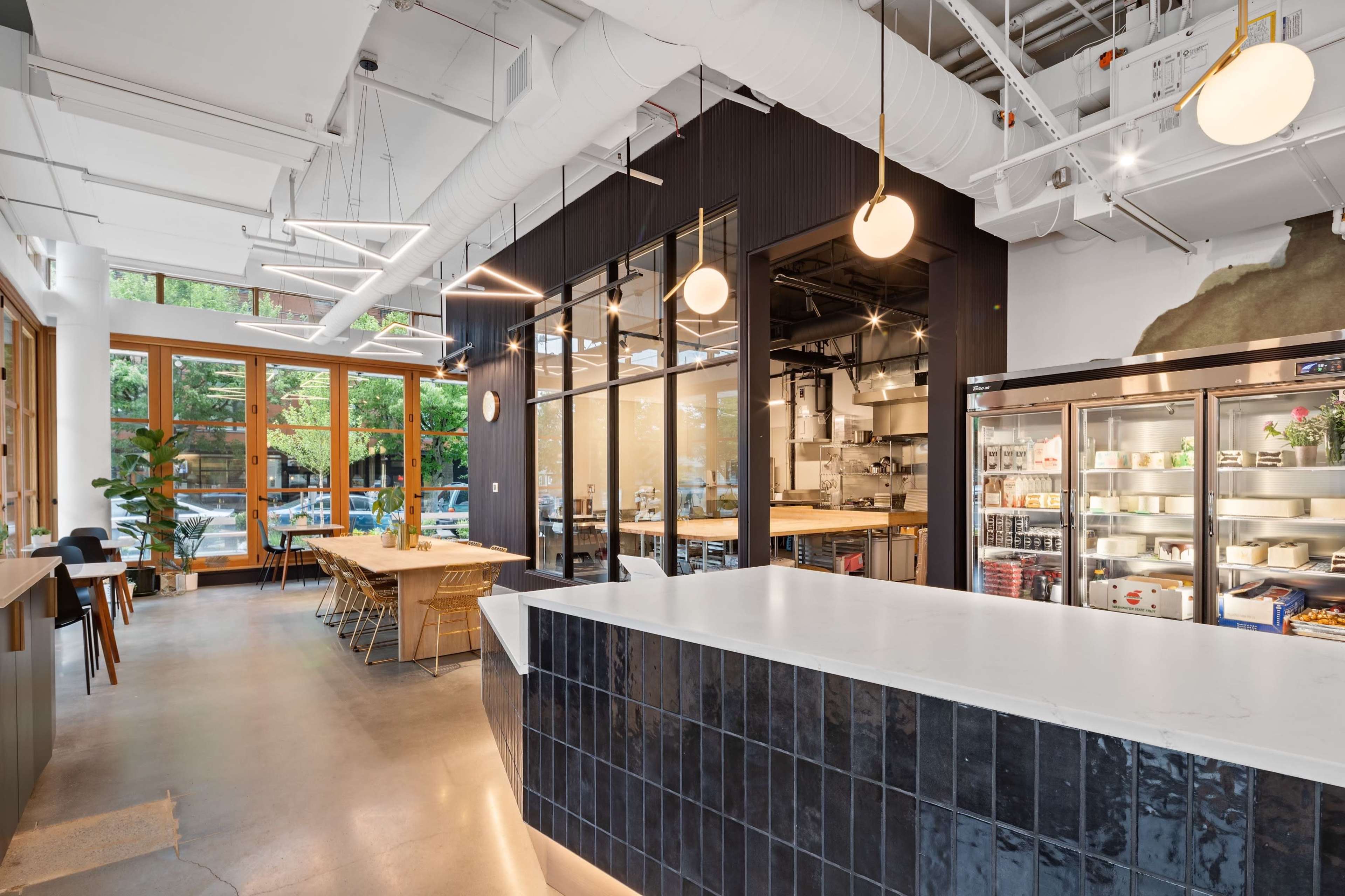 A modern cafe interior with large windows, a long white countertop, wooden tables, and a visible kitchen area in the background.