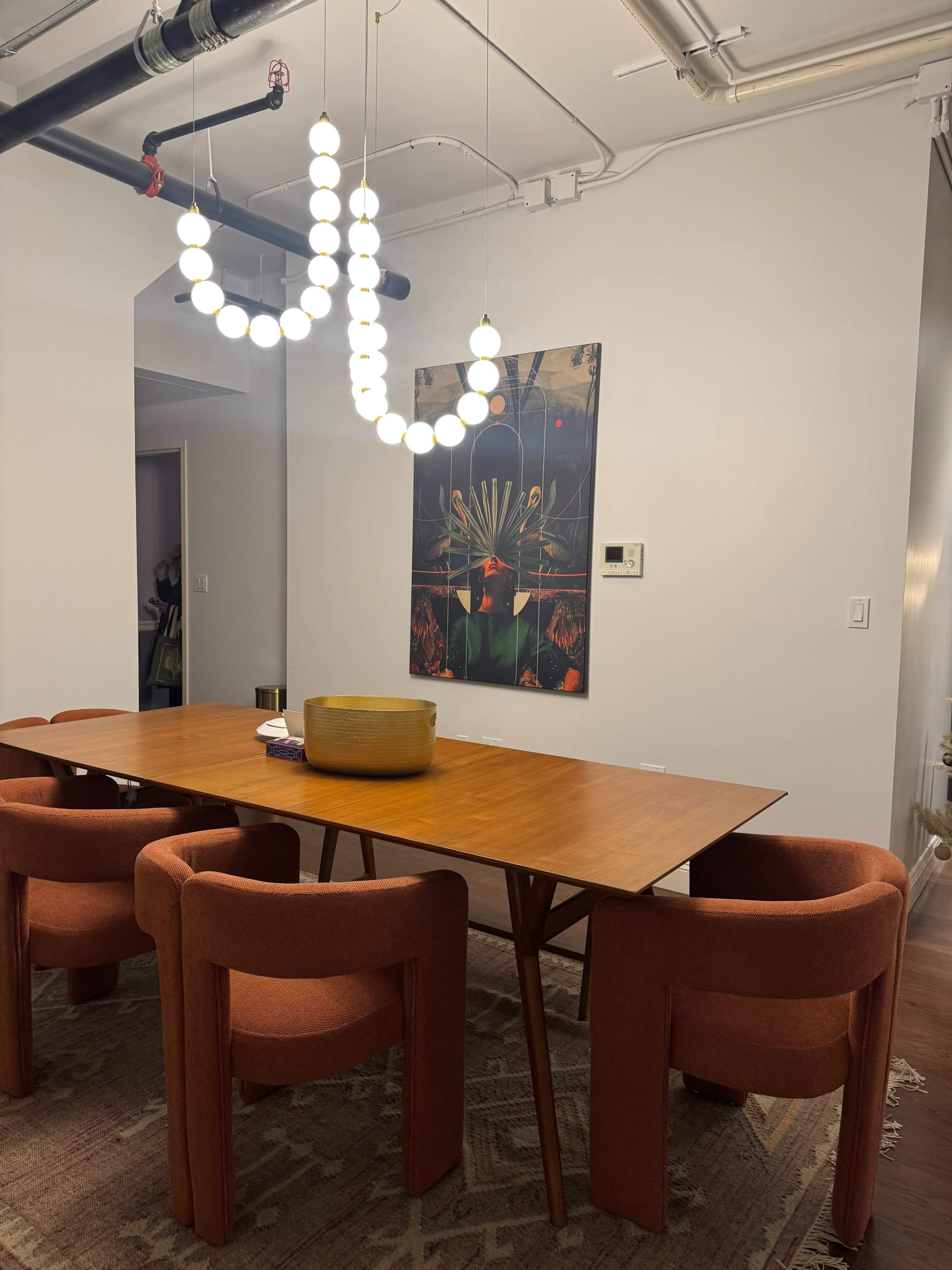 A modern dining area features a wooden table surrounded by four upholstered chairs, with a unique chandelier overhead and a decorative artwork on the wall.