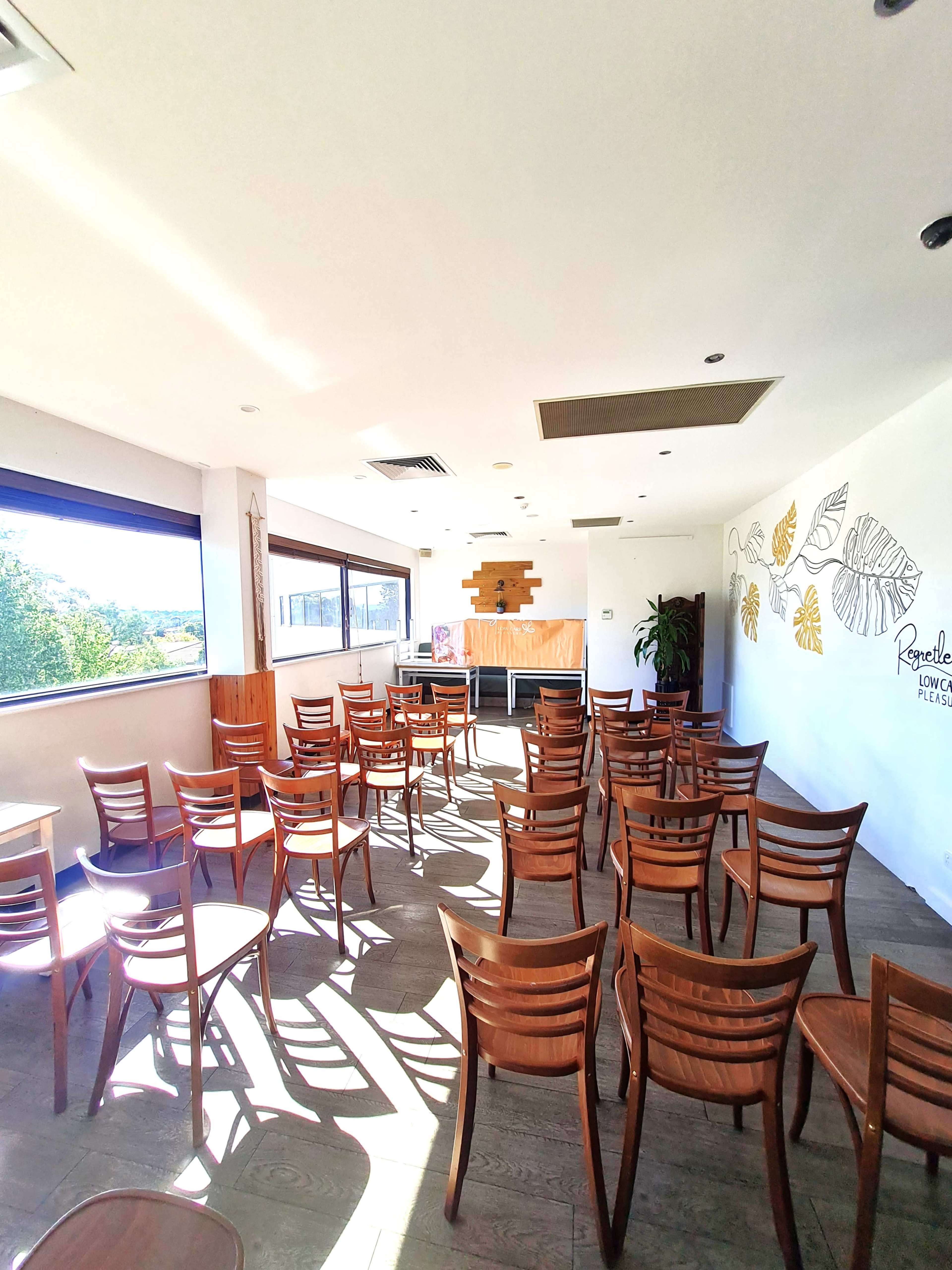 The image shows a bright and spacious room with rows of wooden chairs arranged facing a presentation area, illuminated by natural light from large windows.