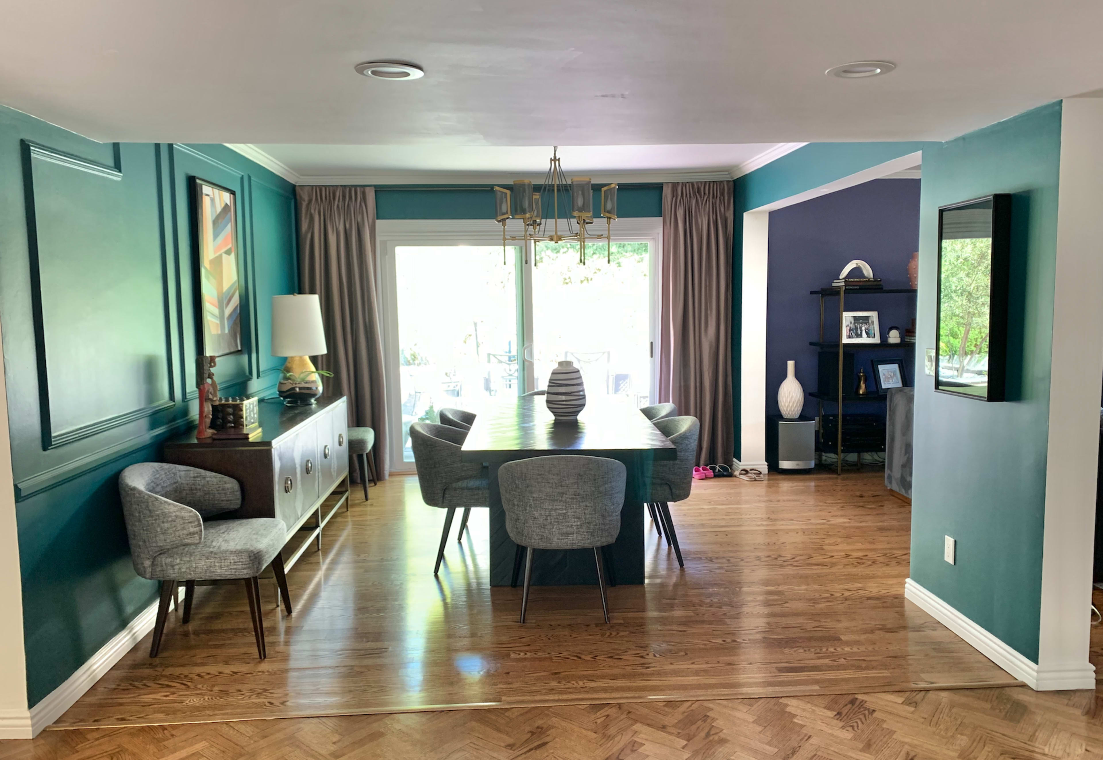 The image shows a dining area with a large table surrounded by six chairs, adjacent to a living space with sliding glass doors leading to an outdoor area.