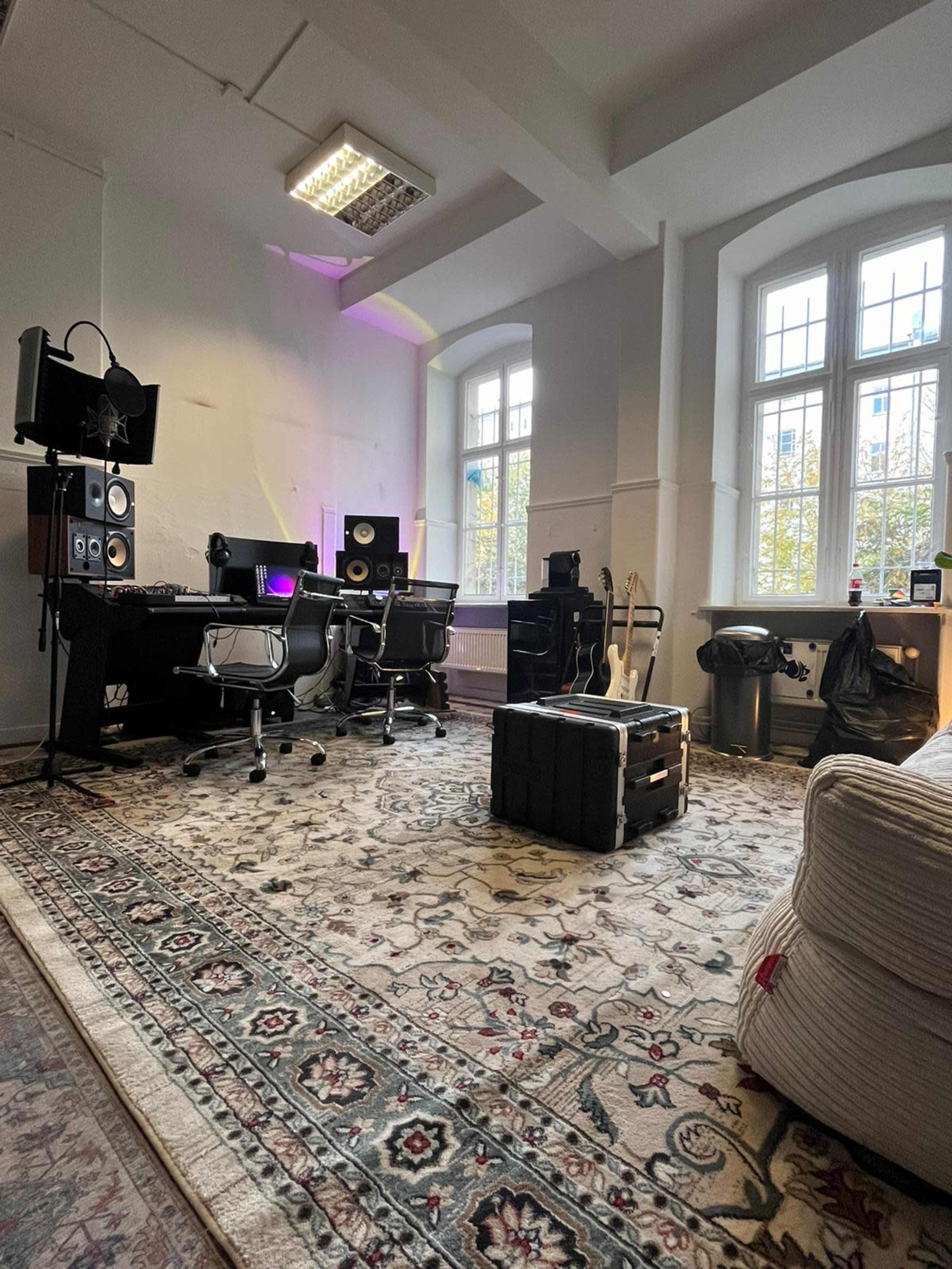 The image shows a music production studio featuring two office chairs, a desk with monitors, a guitar, and a large area rug.