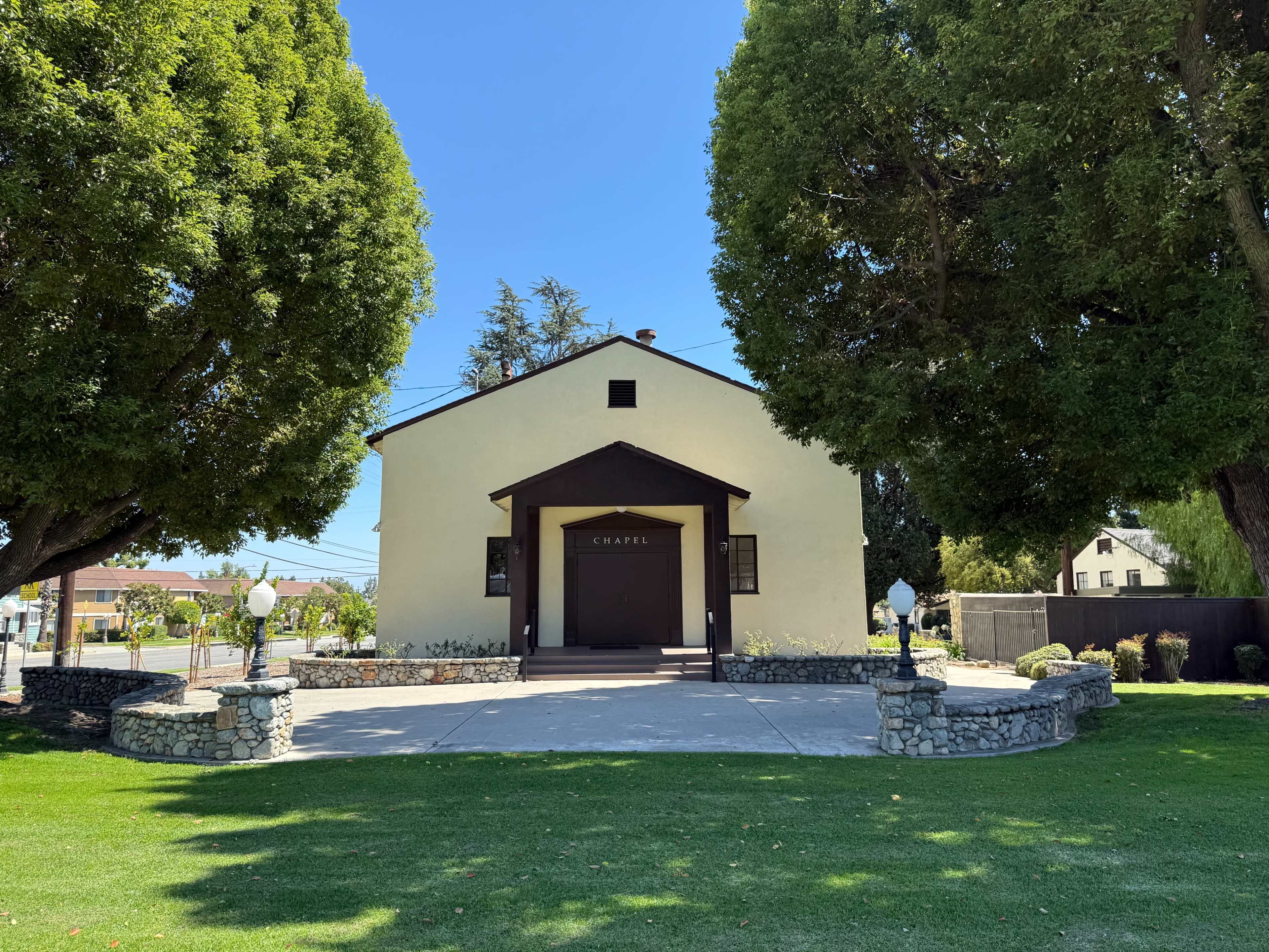 The image shows a chapel with a stone foundation situated on a green lawn, surrounded by large trees.