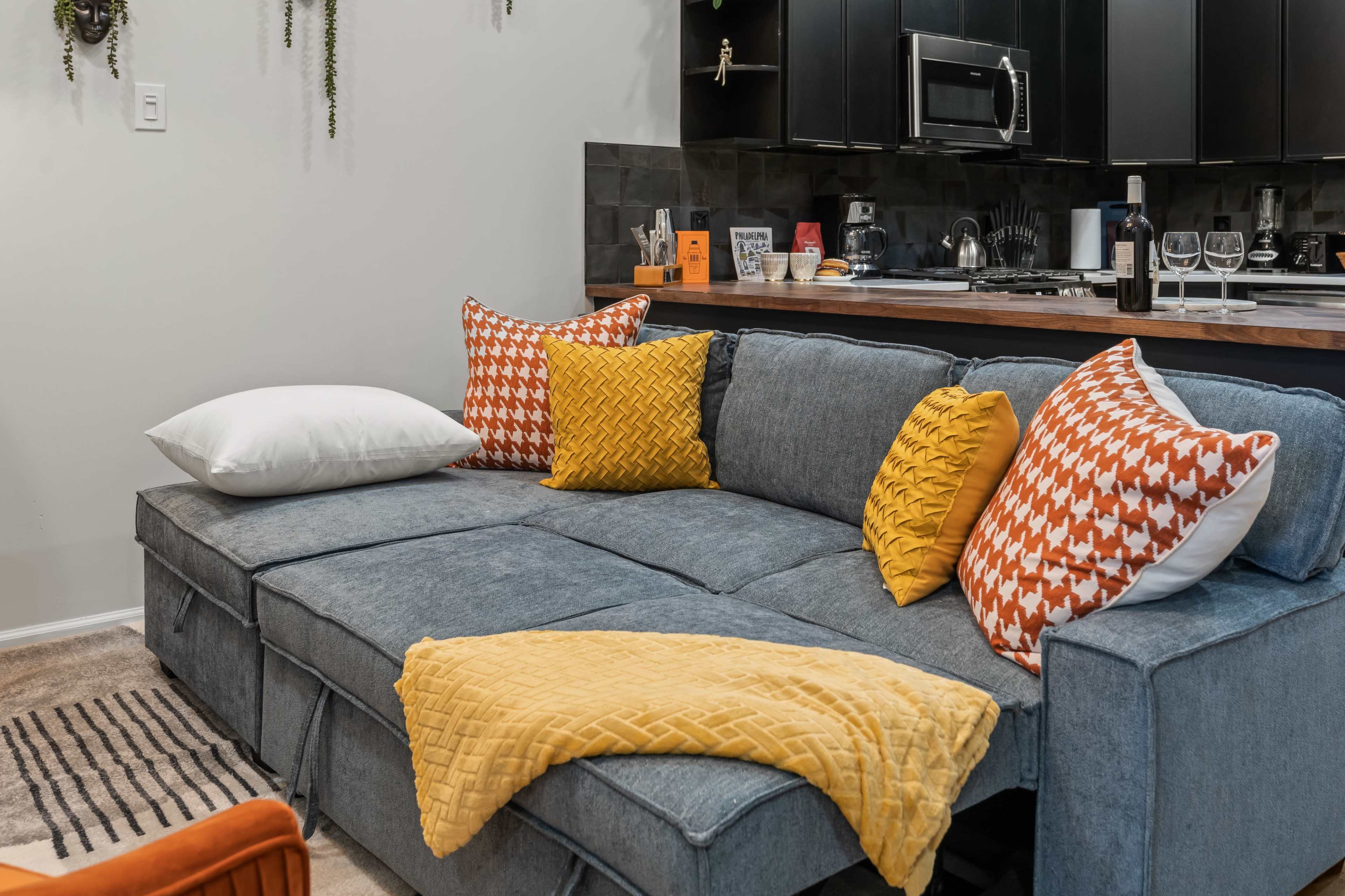 The image shows a comfortable gray sectional sofa adorned with orange and yellow patterned pillows, accompanied by a cozy throw blanket, set in a modern kitchen environment.