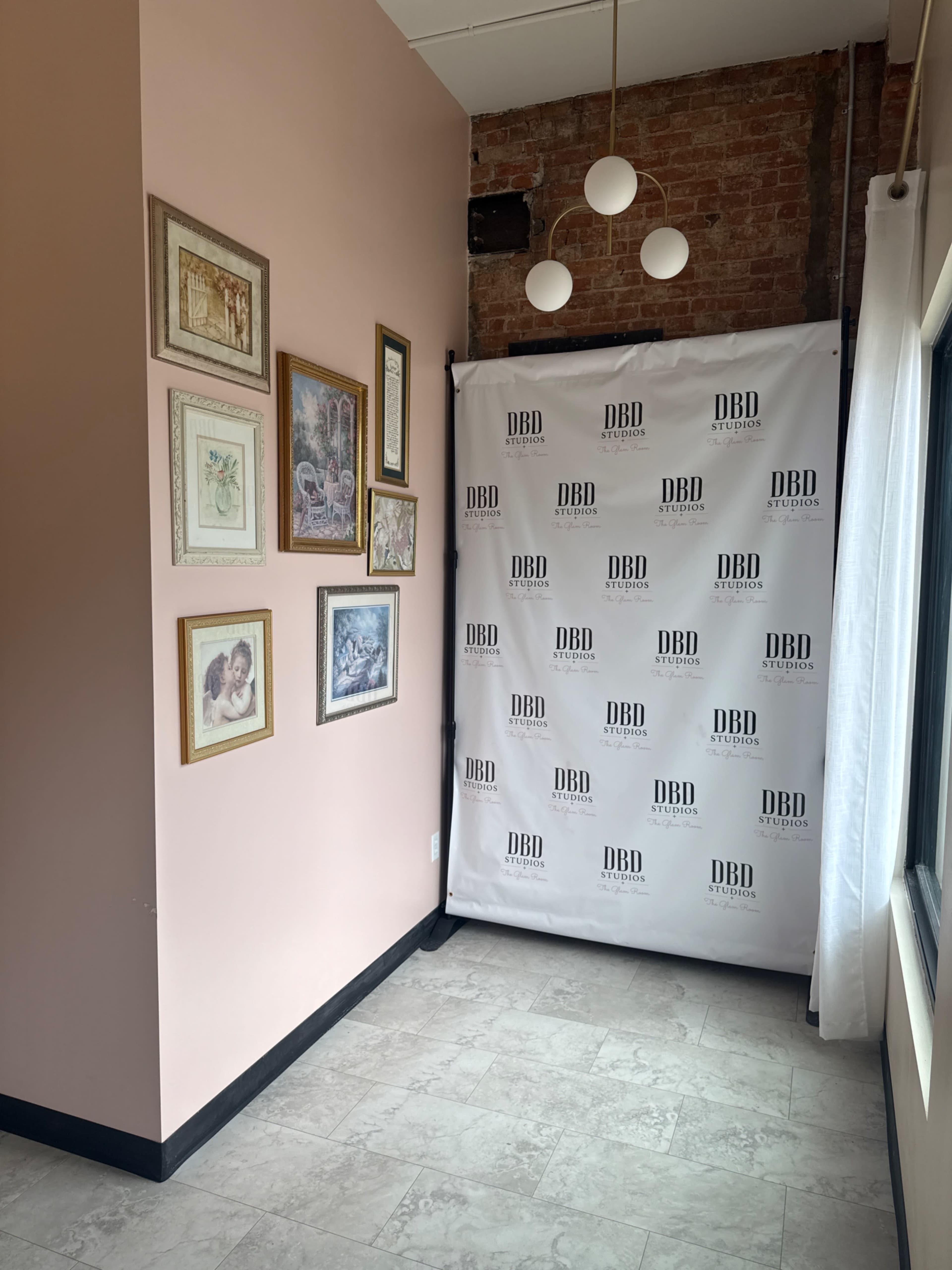 The image shows a corner of a room with a pink wall, a series of framed artworks on one side, and a white backdrop with the text "DBD Studios" on the other.