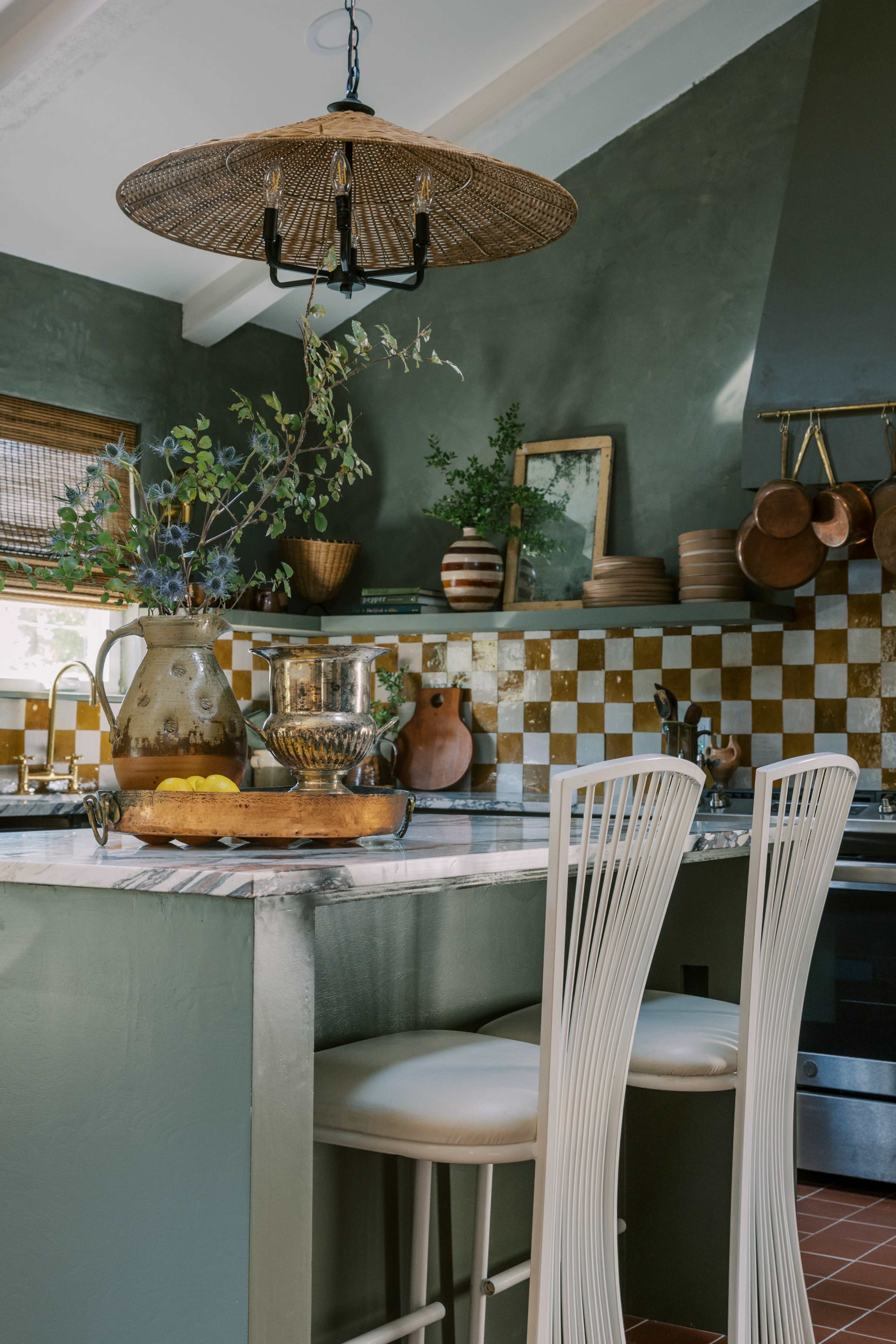 A kitchen features a marble island with seating, dark green walls, woven pendant lighting, and decorative cookware arranged on the countertop and walls.