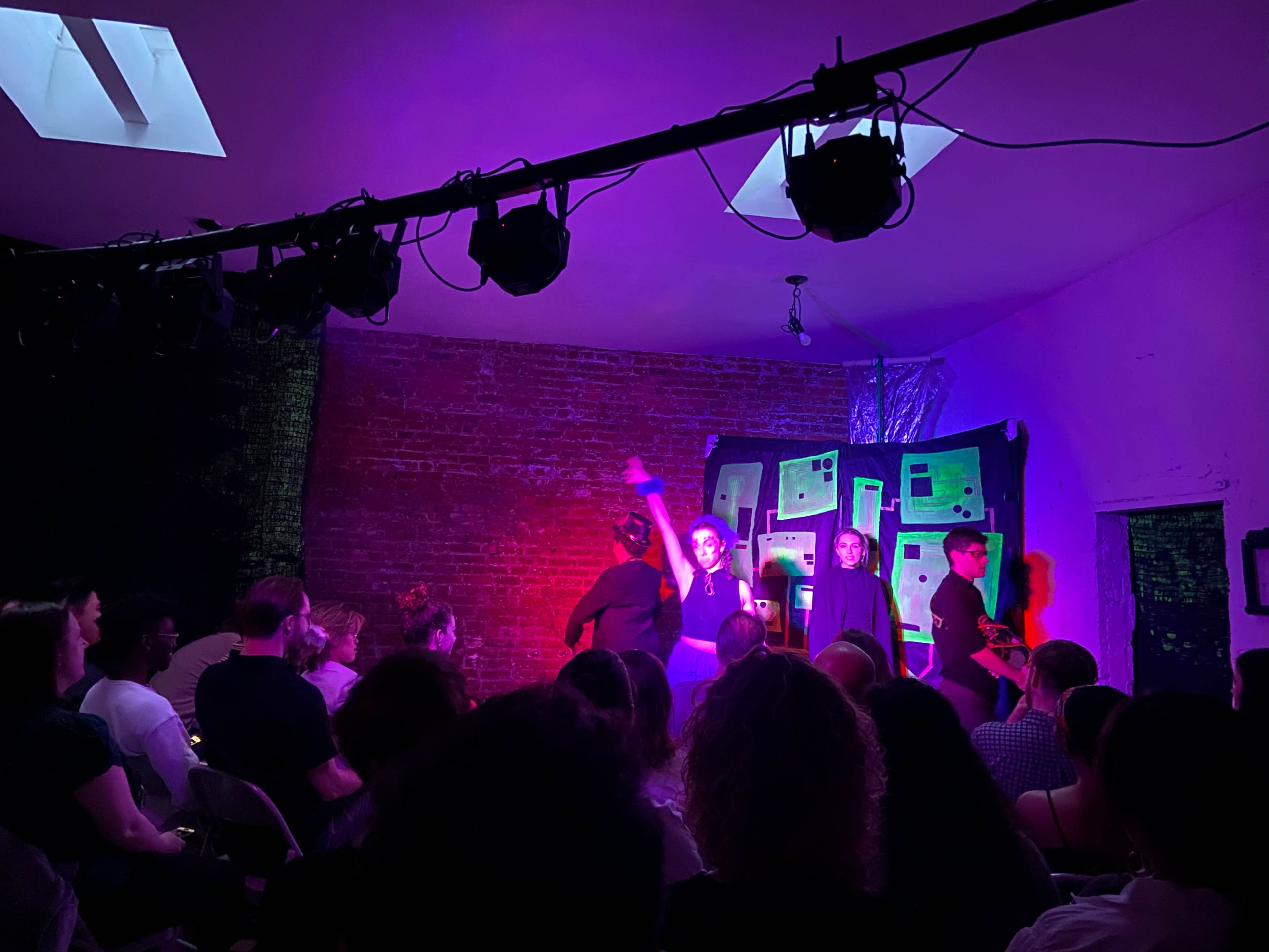 A group of performers is on stage during a theatrical performance, while an audience observes from their seats in a dimly lit room.