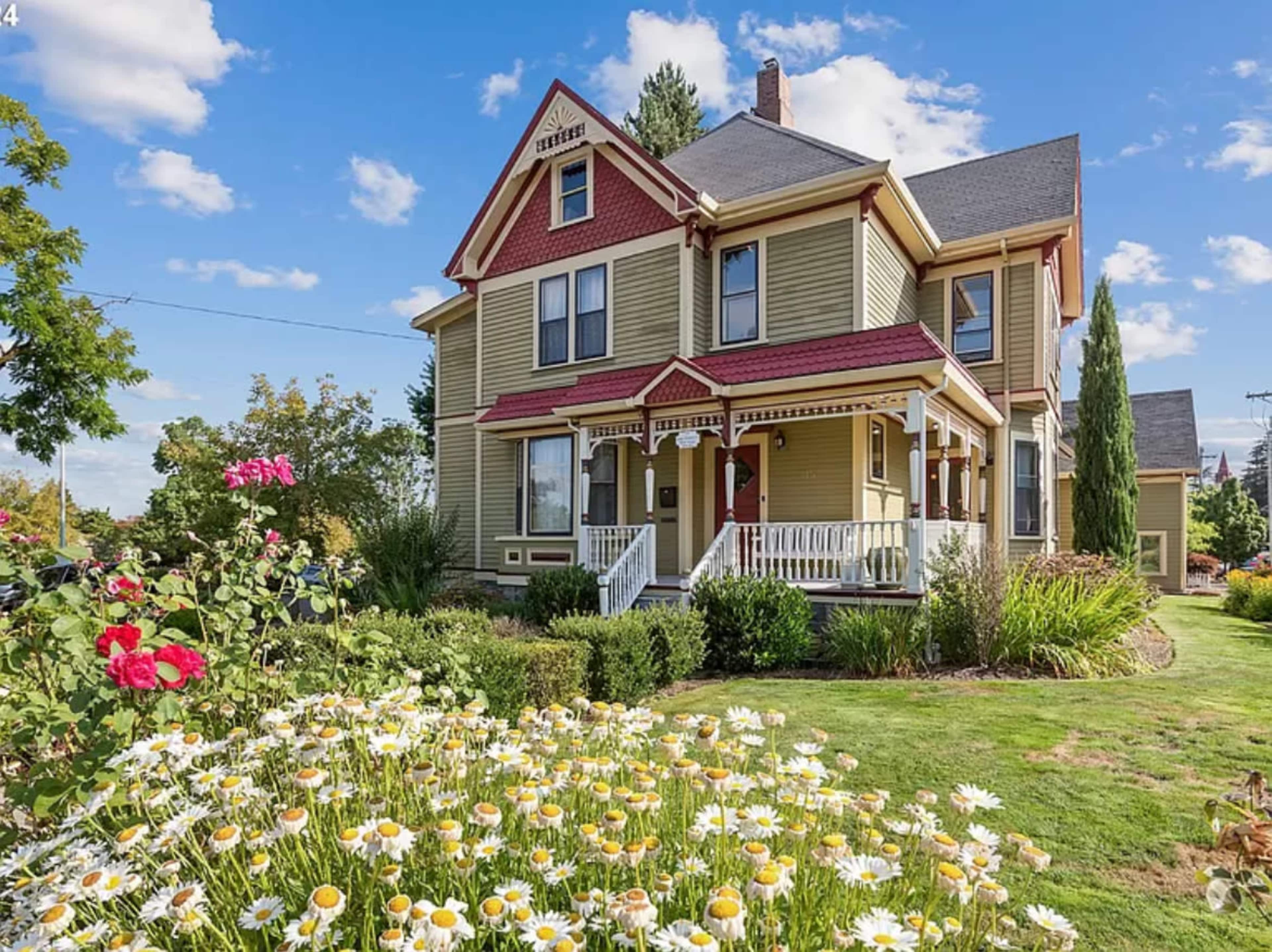 A Victorian-style house with a green exterior and red accents is surrounded by a garden of daisies and roses under a blue sky with scattered clouds.