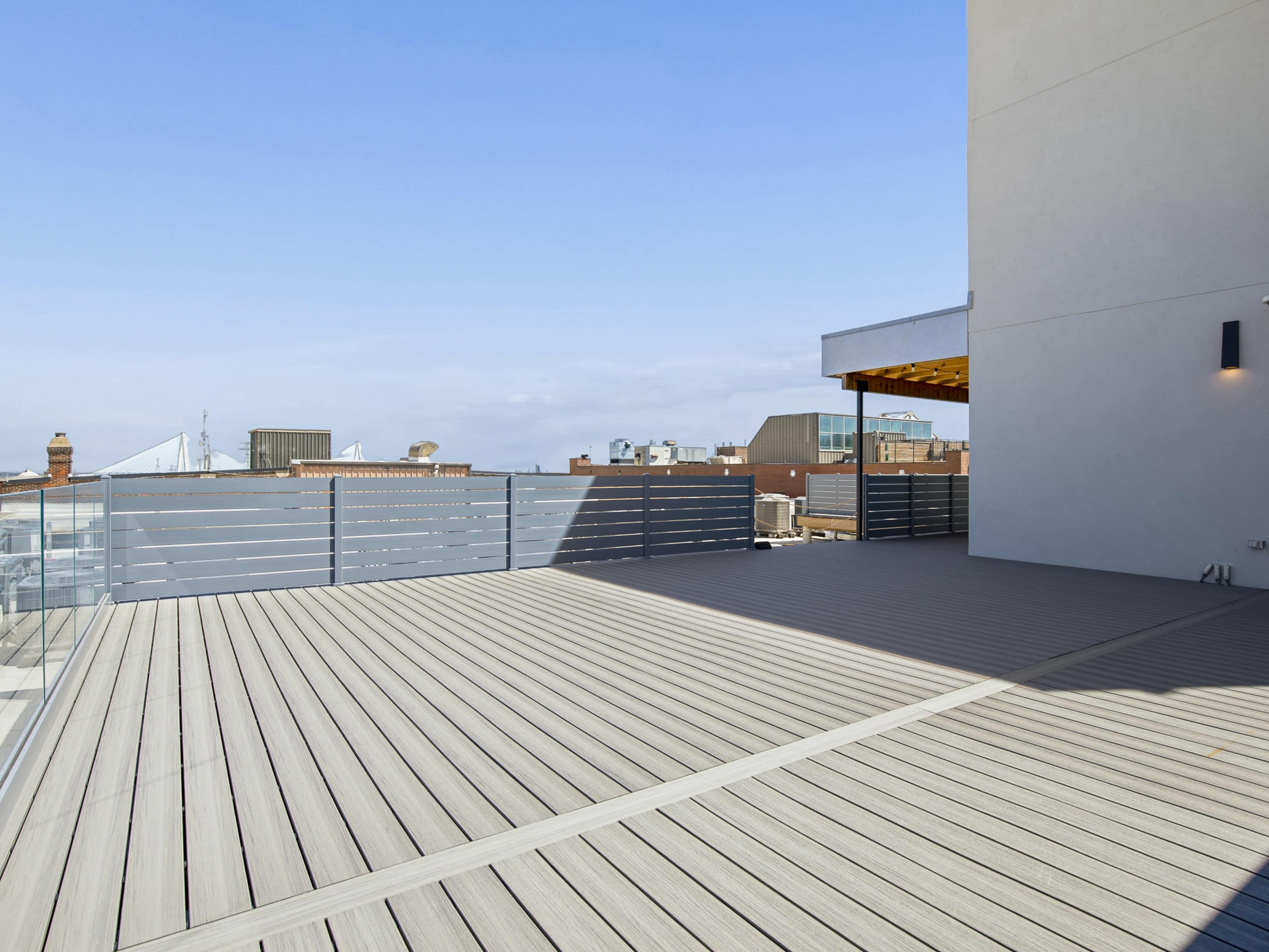 The image shows a spacious wooden deck with a gray railing, overlooking city rooftops under a clear sky.