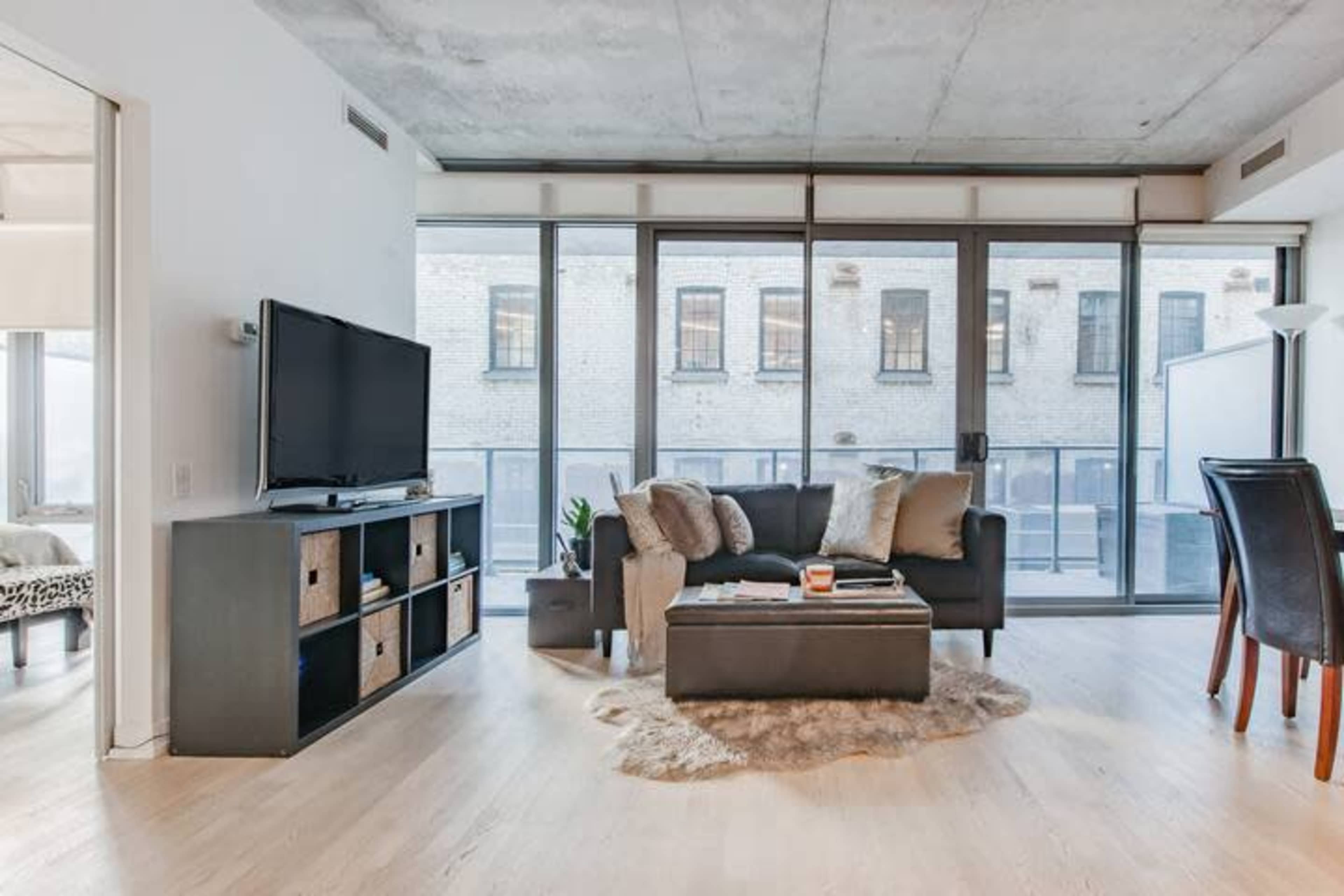 A modern living room featuring a dark couch, a coffee table, and a television, with large windows revealing a brick building outside.