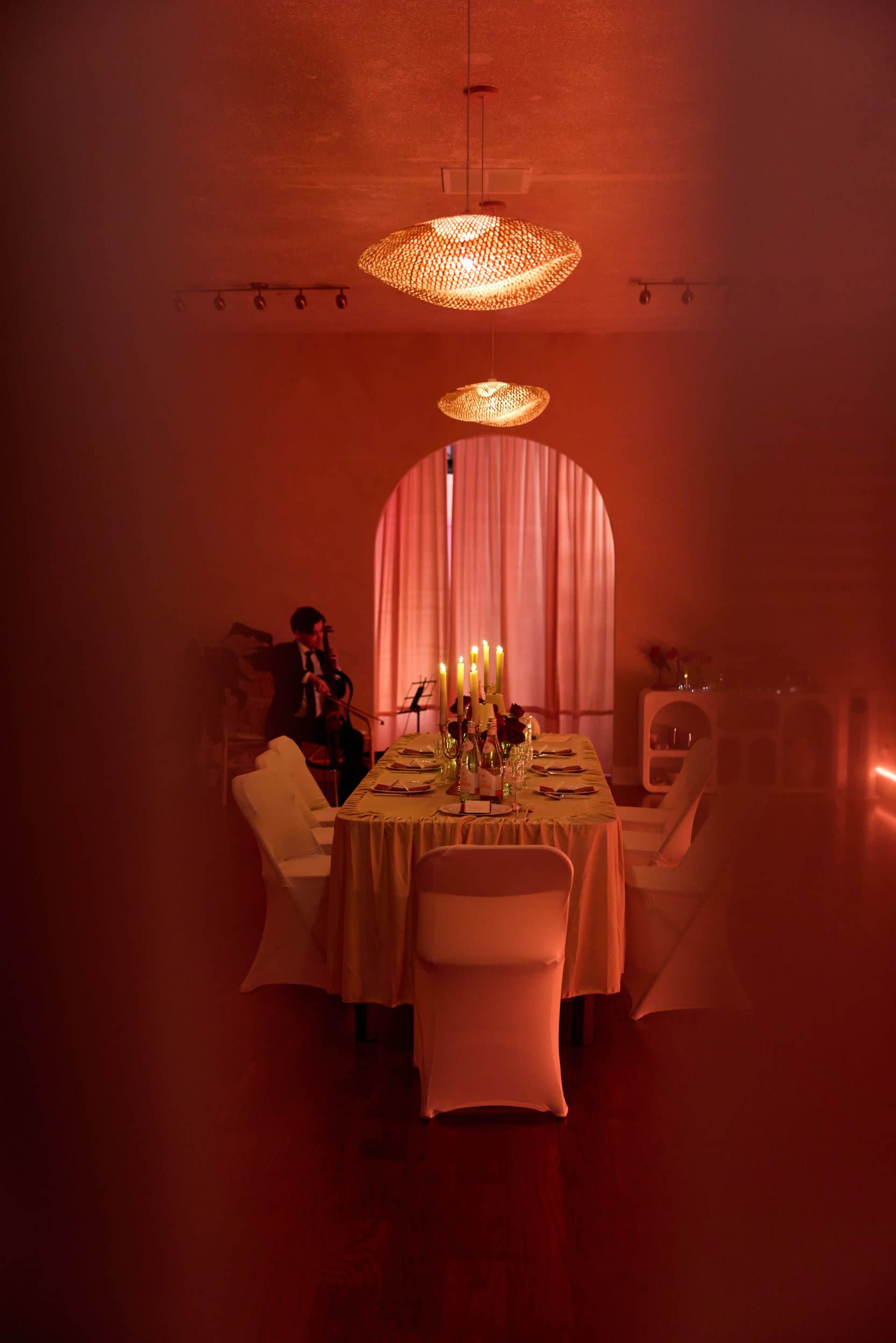 A dining table with candles and flowers is set in a warmly lit room, while a musician plays in the background.